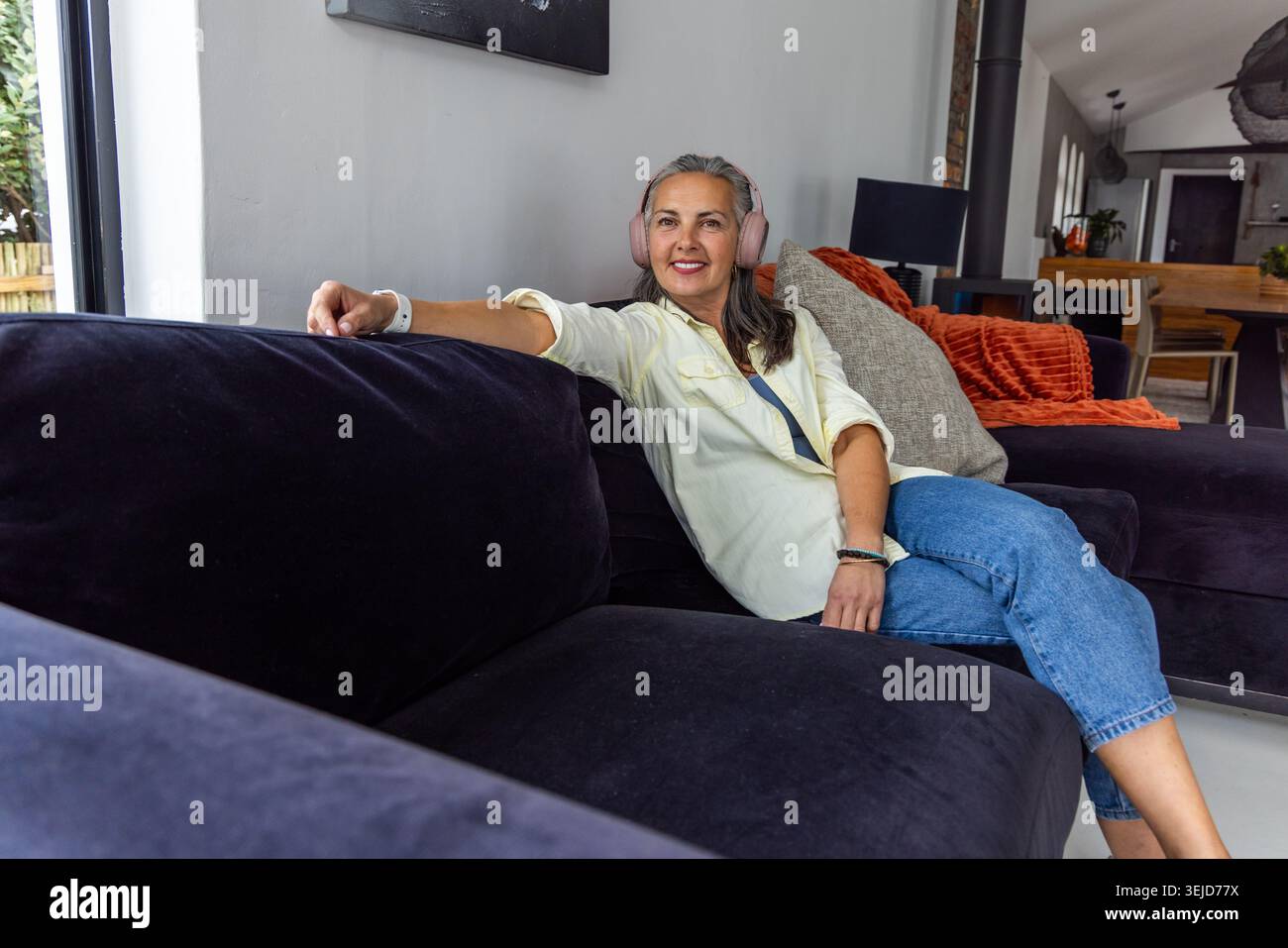 Frau, die sich seitlich auf dunklem Samtschnitt zu Hause entspannt, rosa Kopfhörer trägt und lächelt Stockfoto