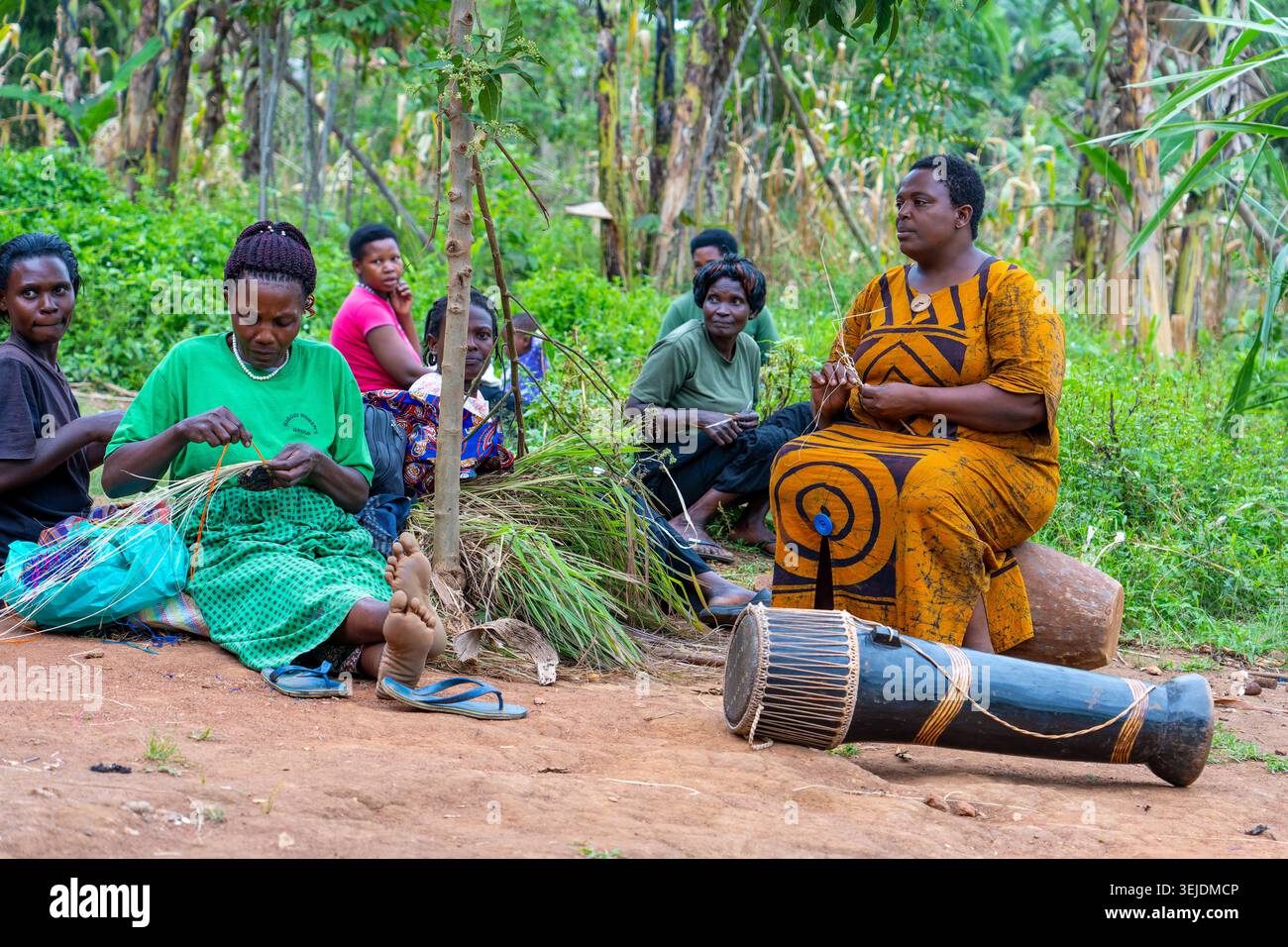 Gruppe afrikanischer Frauen, die auf dem Boden sitzen, lächeln und sich an traditionellem Handwerk des Webenens von Raffiakörben aus natürlichen Materialien beteiligen, umgeben von B Stockfoto