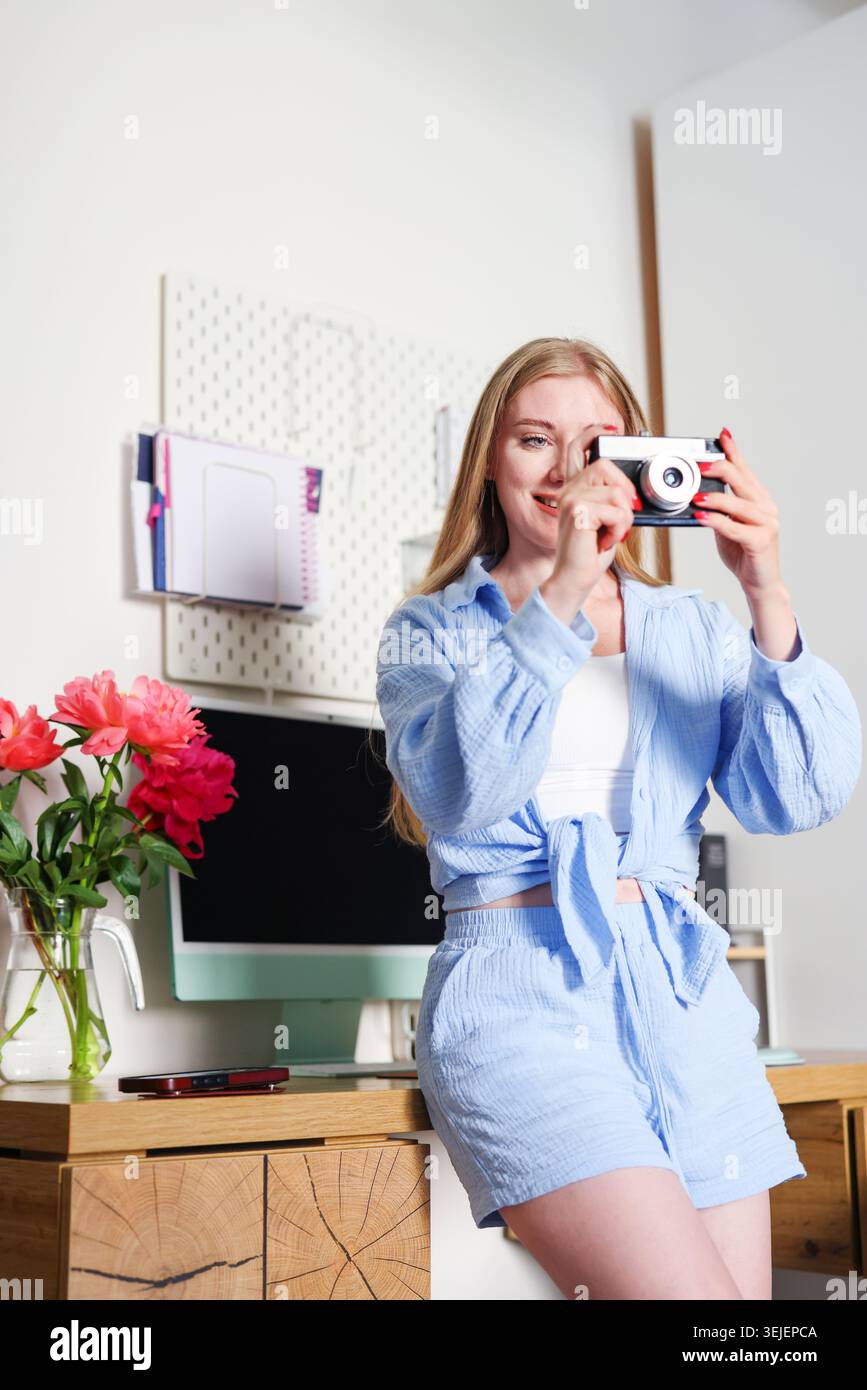 Junge Frau, die Fotos mit Retro-Kamera im gemütlichen Heimbüro macht, kreatives Lifestyle-Porträt mit Blumen, Technologie und Remote-work-Vibe. Stockfoto