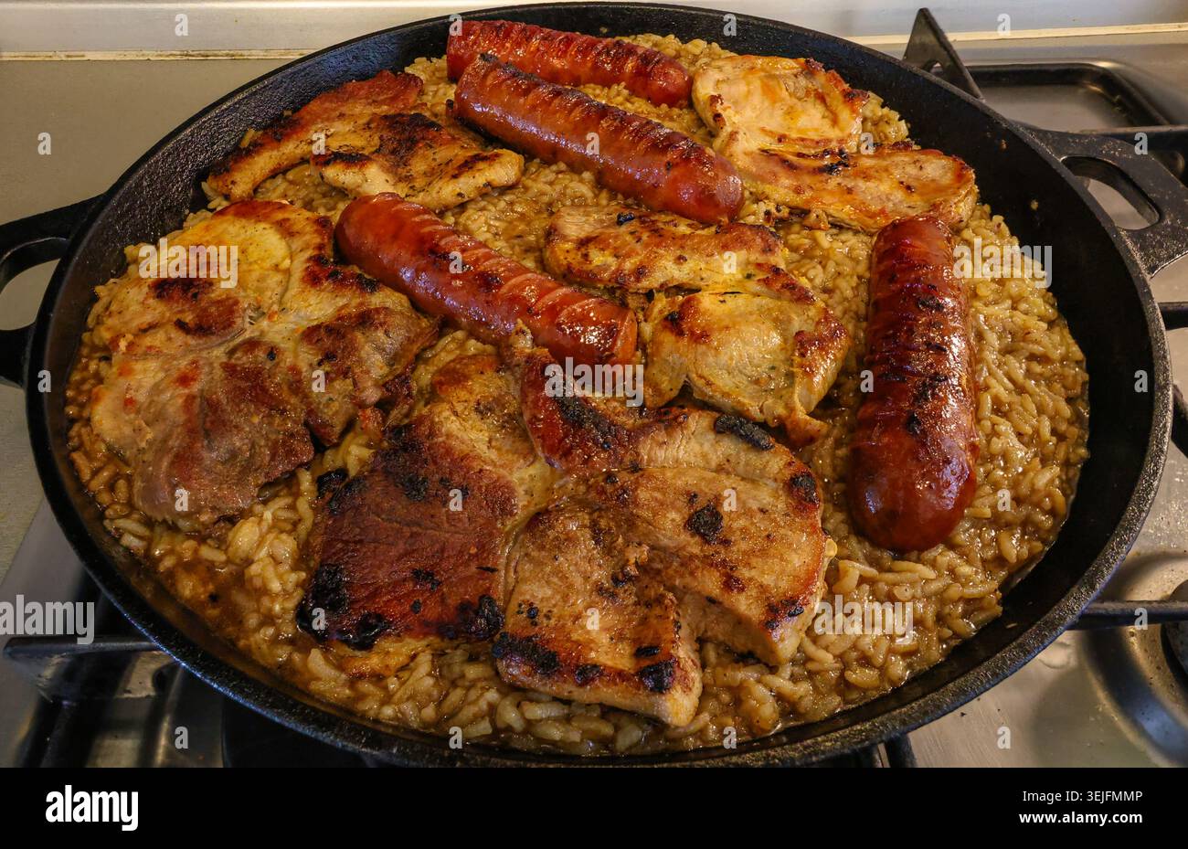 Herzhafte gegrillte Schweinekoteletts und gebräunte Würstchen serviert auf würzigem Reis in einer großen gusseisernen Pfanne, ideal für rustikale Familienmahlzeiten, Komfort Stockfoto