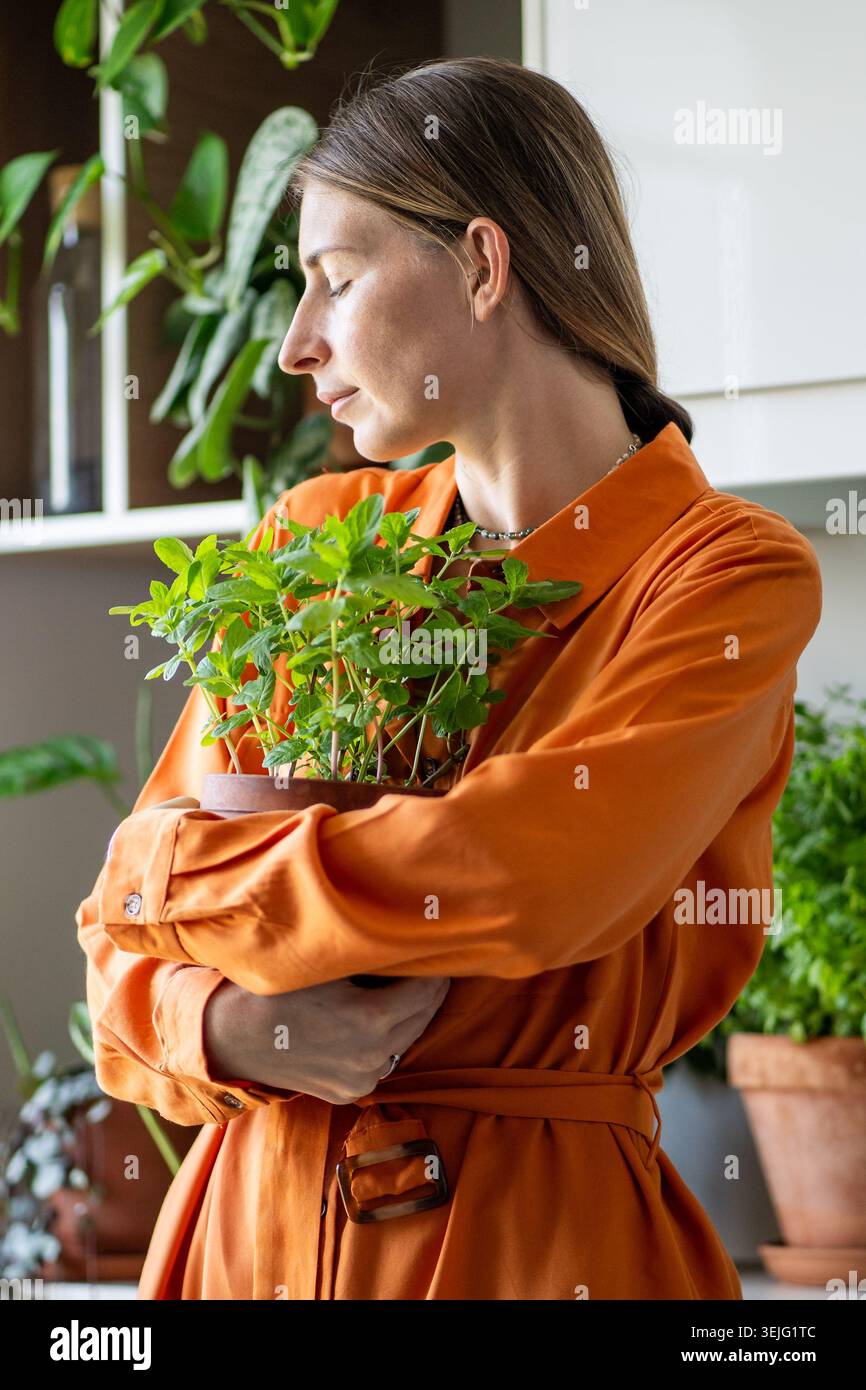 Entspannt lächelnde Frau, die einen Topf mit hausgemachter Pfefferminze in den Händen hält, genießt das Hobby im Innenbereich. Stockfoto