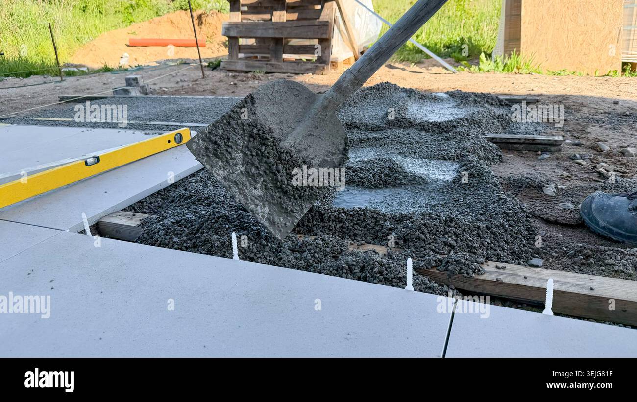 Bauarbeiter, der mit einer Schaufel Betonmischung auf eine Fundamentplatte auf einer Baustelle mit Holzpaletten und Gras im Hintergrund gießt Stockfoto