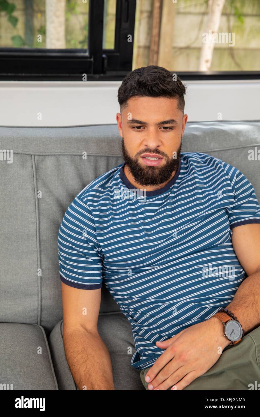 Bärtiger Mann, der auf grauem Sofa unter schwarz gerahmtem Fenster im Wohnzimmer sitzt und Armbanduhr trägt. Zwanglos, Entspannung, Zuhause, Innenraum Stockfoto