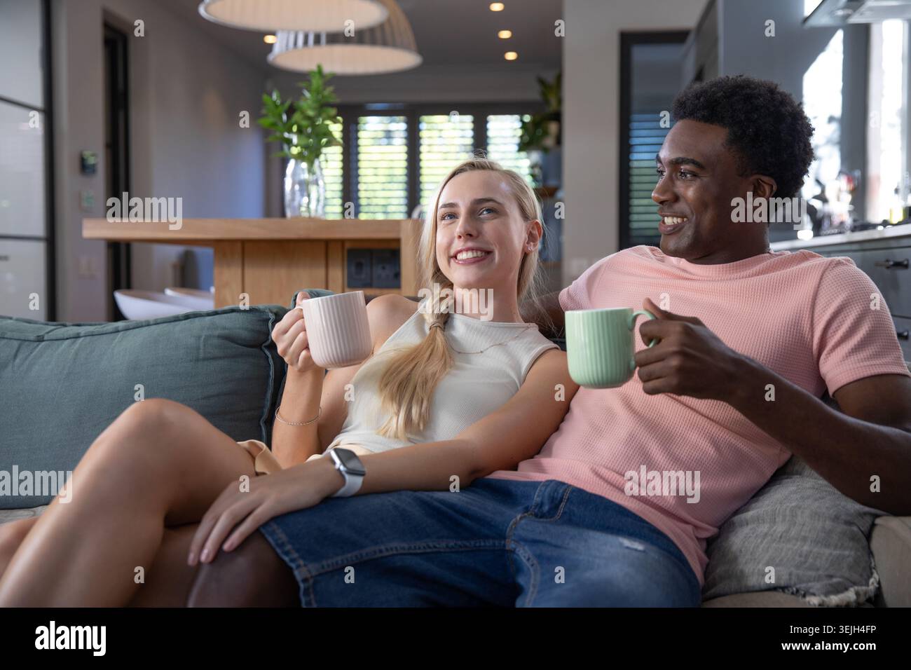 Verschiedene Paare halten Keramikbecher und lehnen sich zurück auf das türkisfarbene Sofa im modernen Wohnbereich. Gemütlichkeit, Komfort, Entspannung, Mod Stockfoto