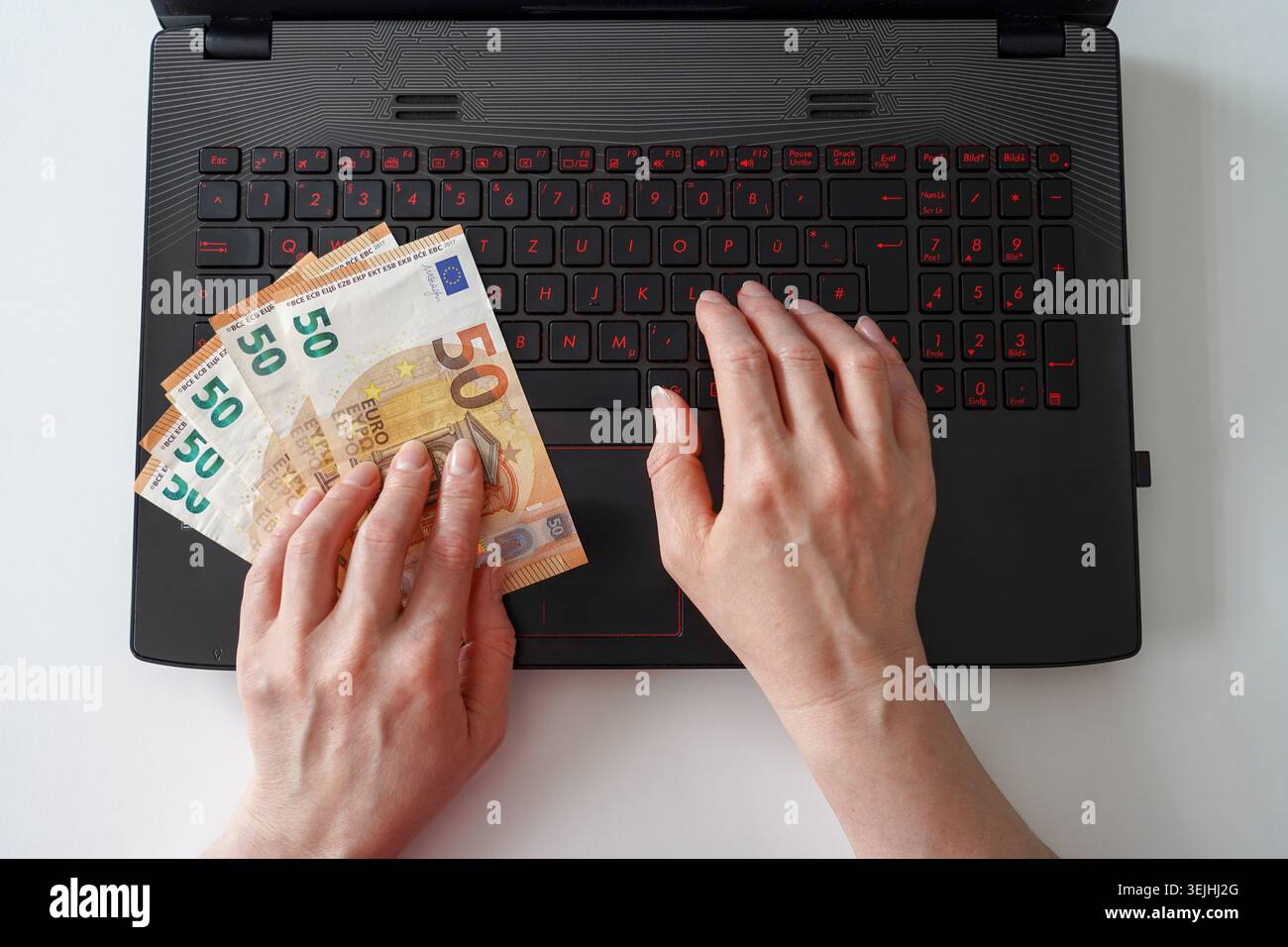 Eine Hochwinkelansicht der Hände, die fünfzig Euro-Scheine halten, während sie auf einer schwarzen Laptop-Tastatur tippen. Stockfoto