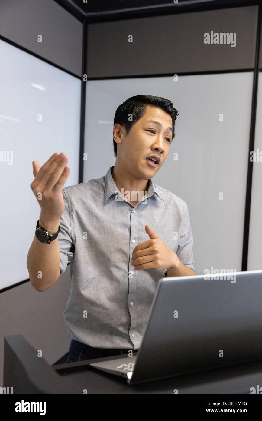 Asiatischer Mann, der hinter dem Podium im Besprechungsraum mit Laptop-Computer und Mattglaspaneelen präsentiert. Professionell, geschäftlich, moder Stockfoto