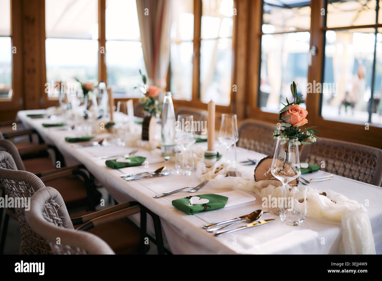 Eleganter Tisch für den Hochzeitsempfang mit weißer Tischdecke, grünen Servietten, Kerzen und frischen pfirsichfarbenen Rosen in rustikaler Holzeinrichtung. Stockfoto