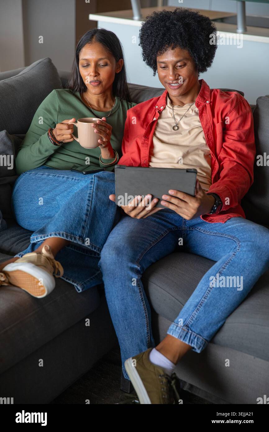 Indische und afroamerikanische Paare sitzen auf einem grauen Sofa im Wohnzimmer und halten ein Tablet und einen Becher. Modern, gemütlich, Lifestyle, mi Stockfoto