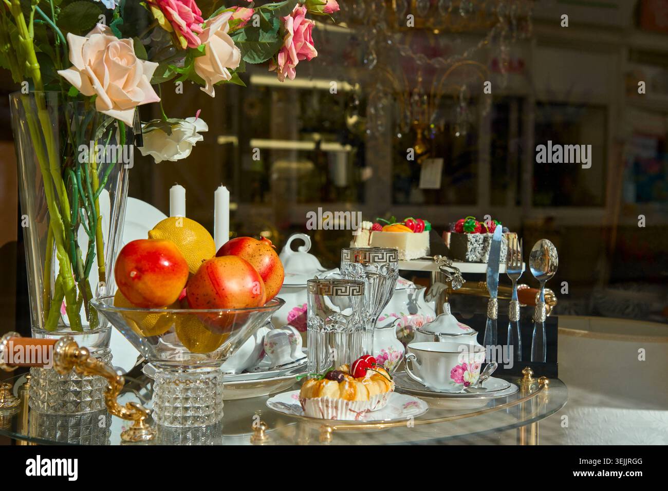 Eleganter Nachmittagsteetisch mit frischem Obst, Kuchen, Kristallgläsern und Rosen hinter einem Schaufenster. Stockfoto