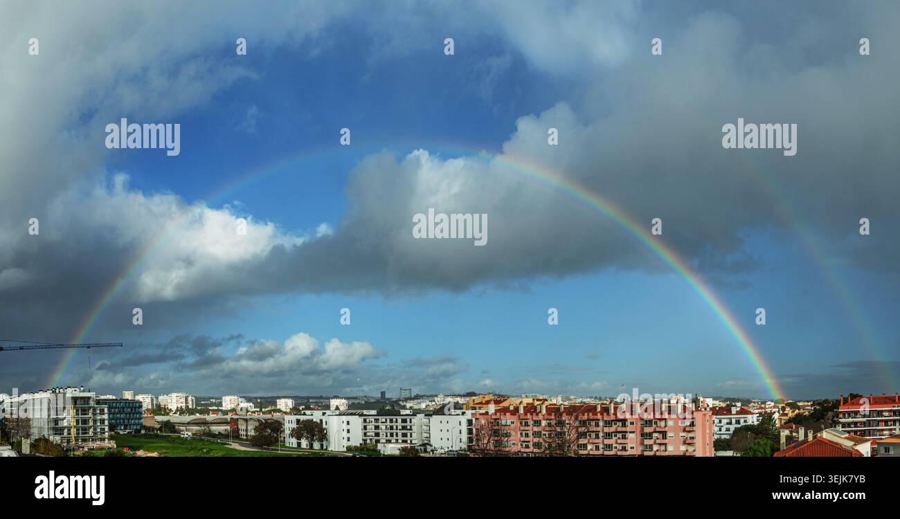 Panoramablick auf einen natürlichen doppelten Regenbogen, der sich nach Regen über einem Wohngebiet erhebt, mit dramatischen Wolken und Sonnenlicht am Himmel. Eine seltene Natu Stockfoto