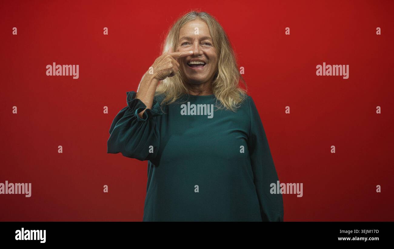Die Seniorin zeigt Finger an Nase und lacht in grünem Kleid vor rotem Studio-Hintergrund; verspielte Freude. Stockfoto