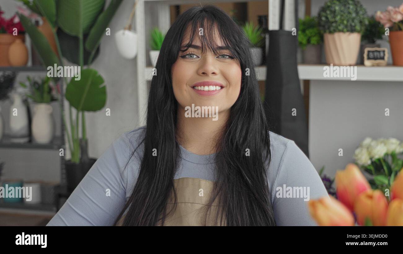 Floristin in Schürze lächelt neben Blumen auf einem Ladentisch im Atelier in die Kamera, mit Topfpflanzen und Vasen herum; Pflanzenpflege pflegt Freude. Stockfoto