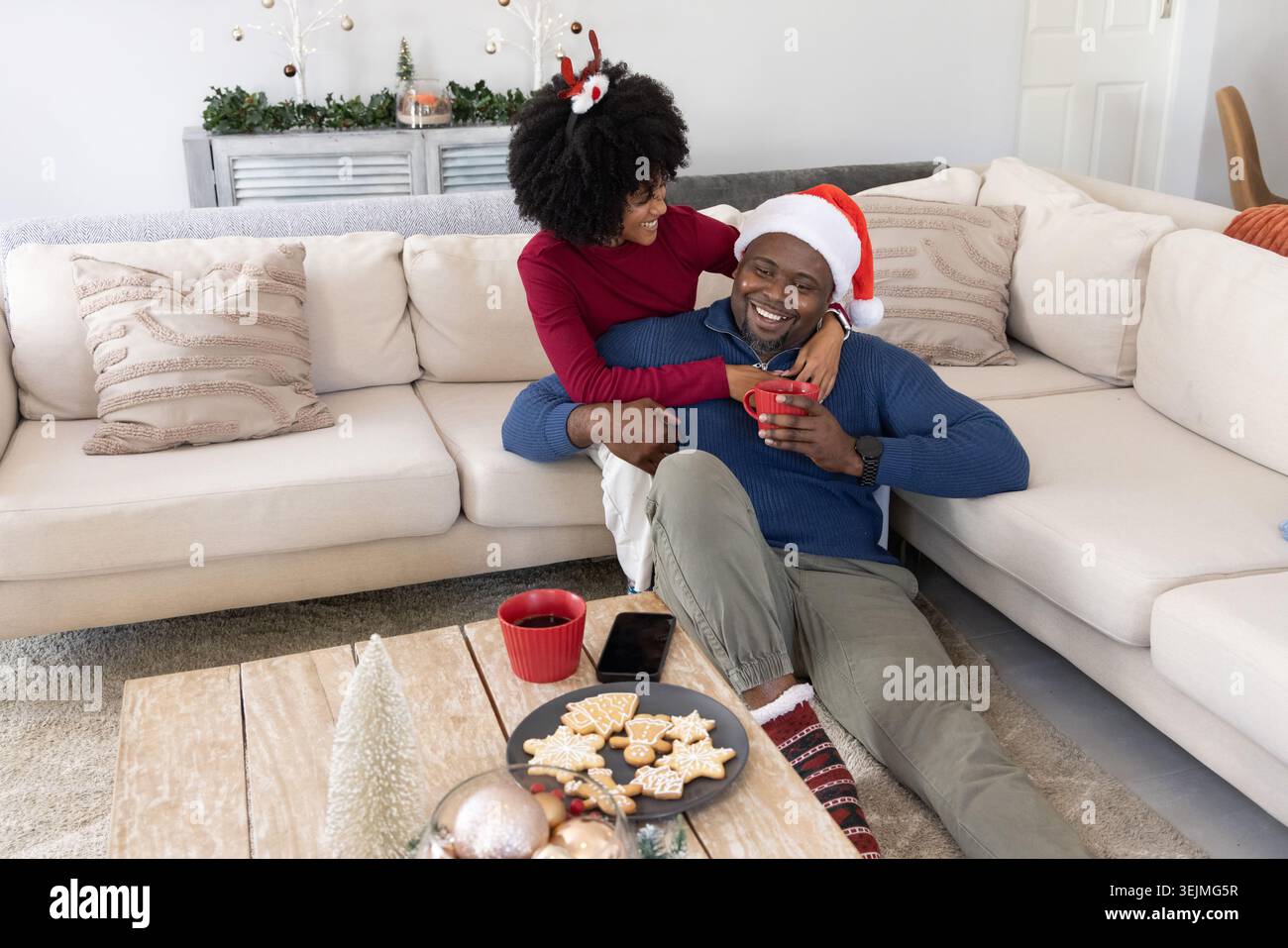 Afroamerikanisches Paar kuschelt zu Hause, Frau im Geweih, Mann im Weihnachtsmann, Kekse auf dem Tisch. Gemütlich, festlich, warm, romantisch, Stockfoto