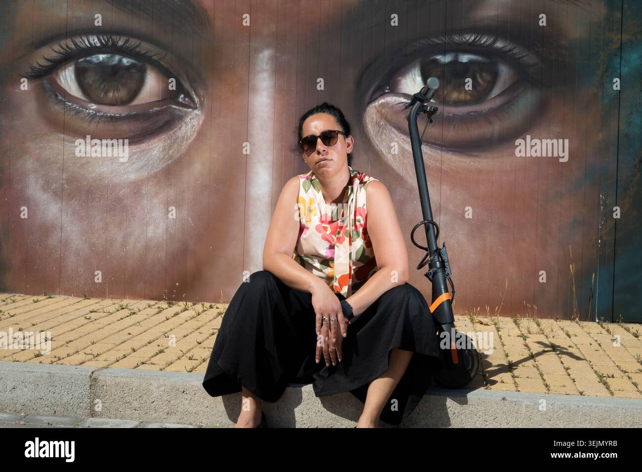 Erwachsene Frau ruht mit Elektroroller an großen Augen Wandgemälde in urbaner Umgebung Stockfoto