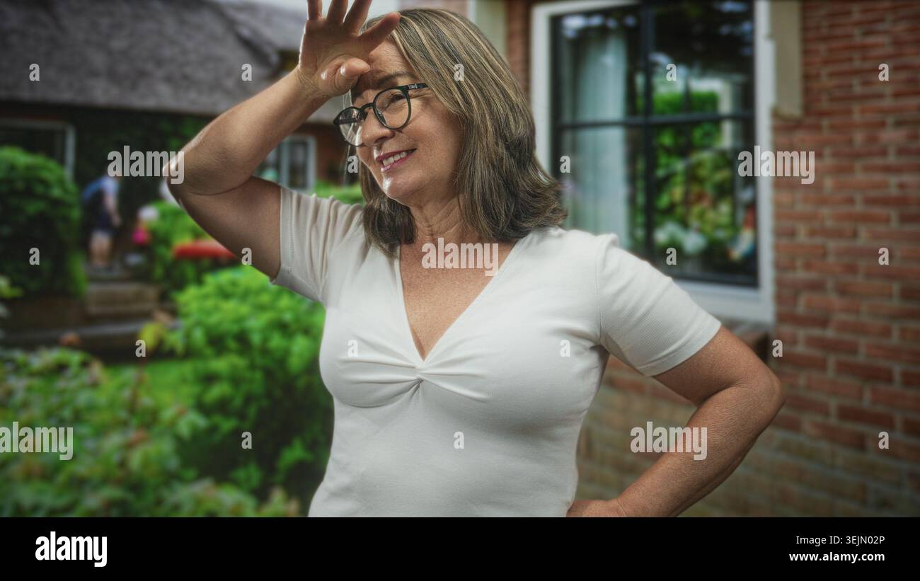 Hispanische Seniorin mit grauem Haar und Brille lächelt, Hände auf Hüften, hebt dann Hand an Stirn, um die Augen zu schattigen, während sie vor einem Gebäude steht; Co Stockfoto