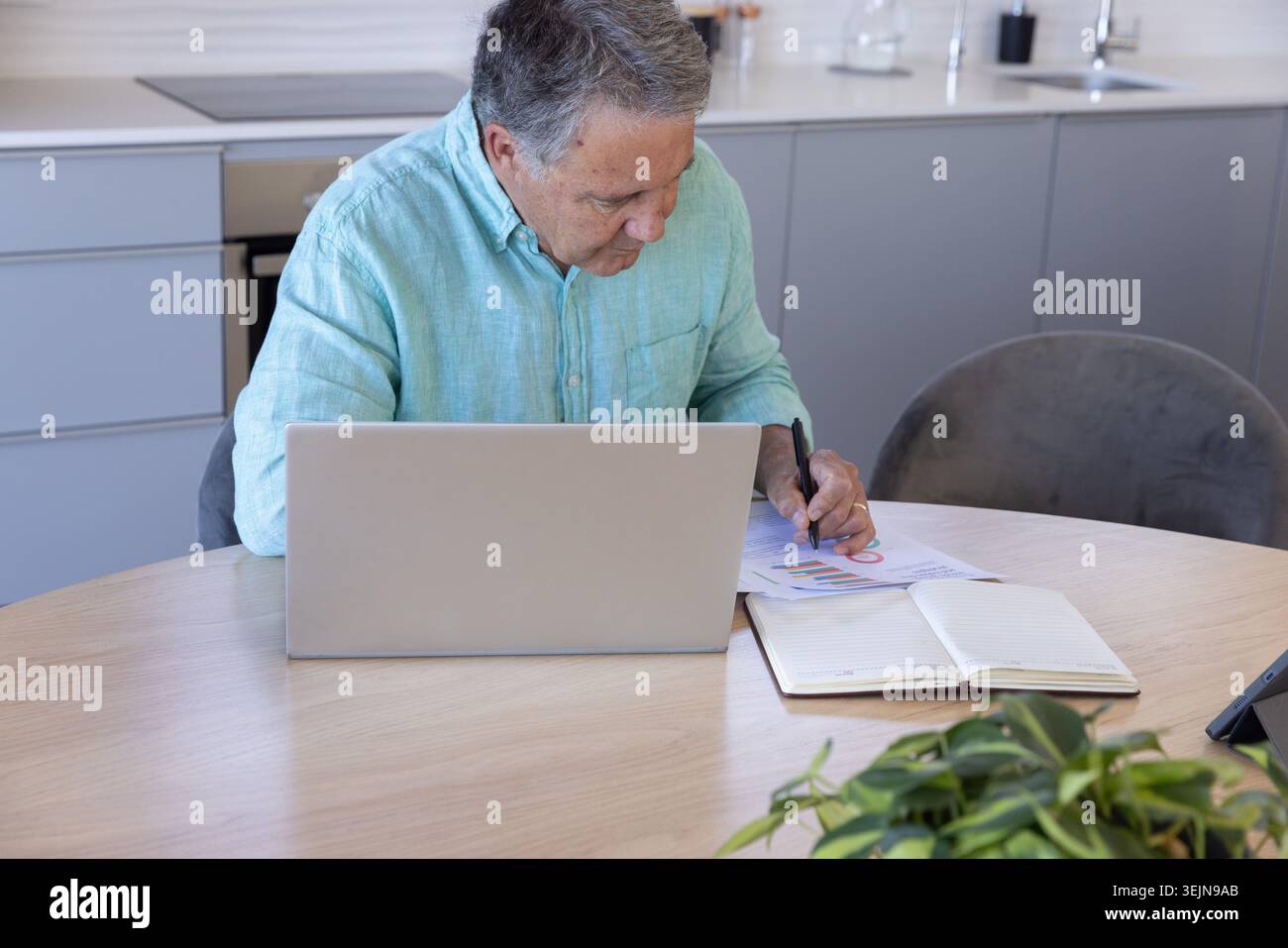 Silberner Laptop, der offen auf einem runden hölzernen Küchentisch mit gedruckten Karten und grüner Pflanze sitzt. HomeOffice, Senior, Finanzen, Plan Stockfoto