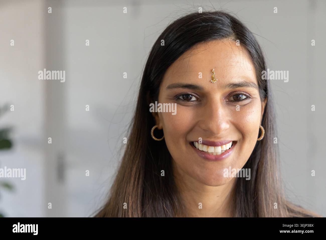 Lächelnde Indianerin posiert zu Hause mit Stirnschmuck, goldenen Ohrringen, Topfpflanzen. Kopfschuss, weiblich, Bindi, Schmuck, in Stockfoto