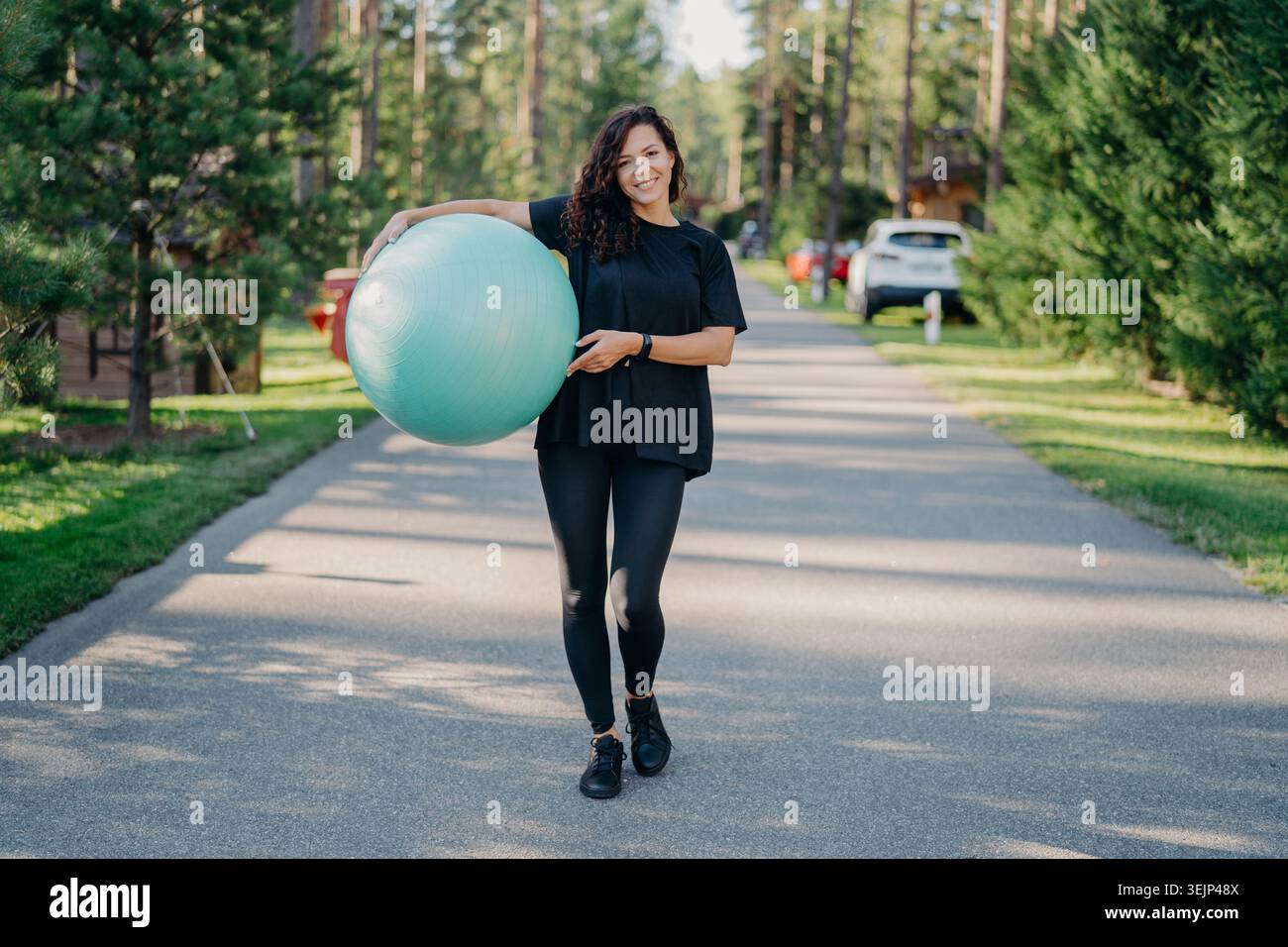 Junge fit Brünette Frau hält großen Fitnessball, trägt aktive Kleidung, macht Workout, posiert im Park, spaziert im Freien und Stockfoto