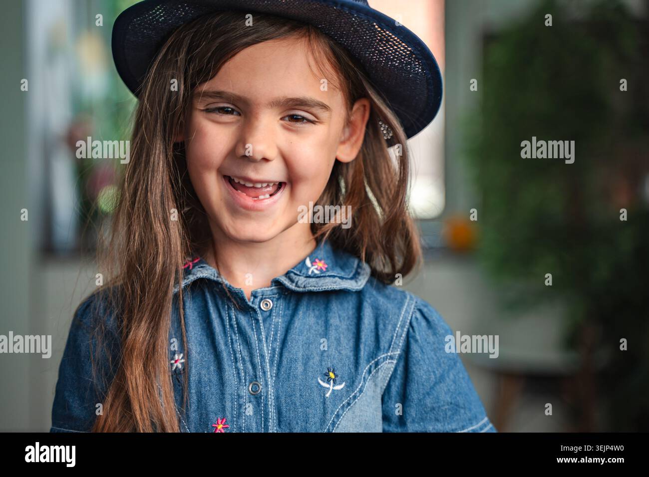 Nahaufnahme eines fröhlichen Mädchens im Vorschulalter, das einen Hut und ein Jeanskleid trägt, in die Kamera blickt und lächelt Stockfoto