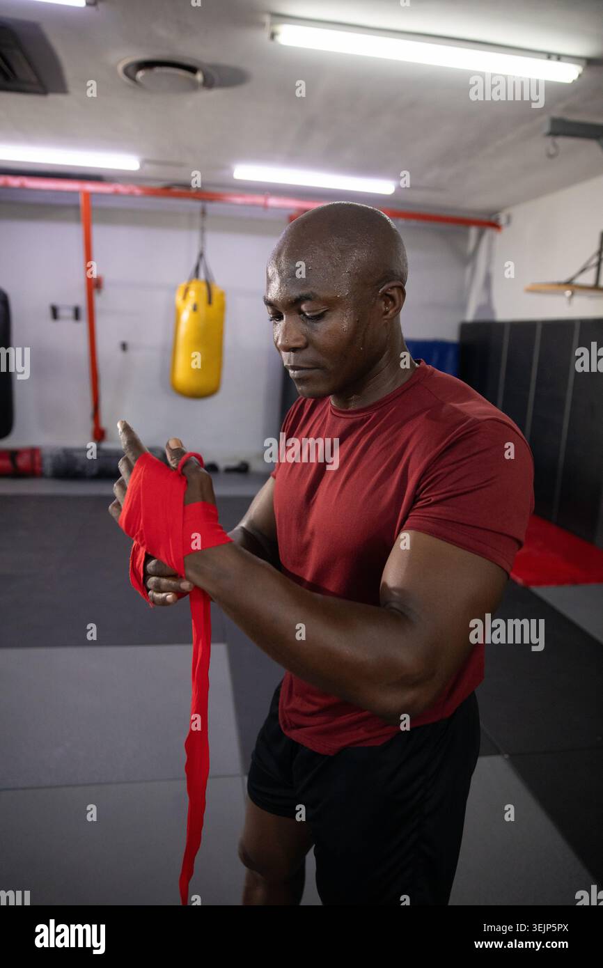 Afroamerikanischer Mann in rotem Hemd, schwarze Shorts, die rote Handhüllen auf Turnmatten in der Nähe der gelben Tasche wickeln. Boxen, Fitnessstudio, Fokus, Schweiß, f Stockfoto