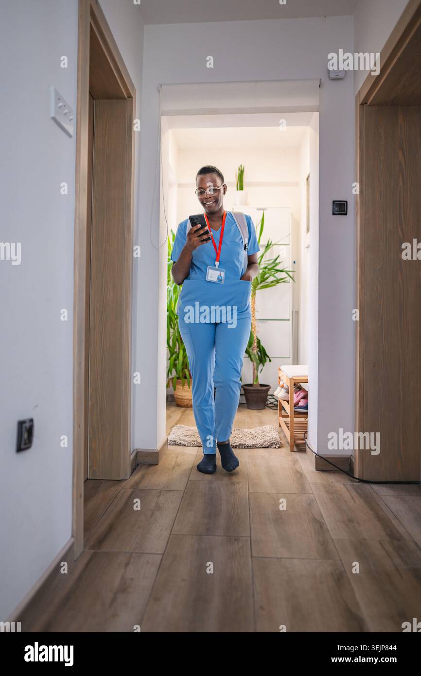 Junge Ärztin in blauem Scrubs mit ihrem Smartphone zu Hause Stockfoto