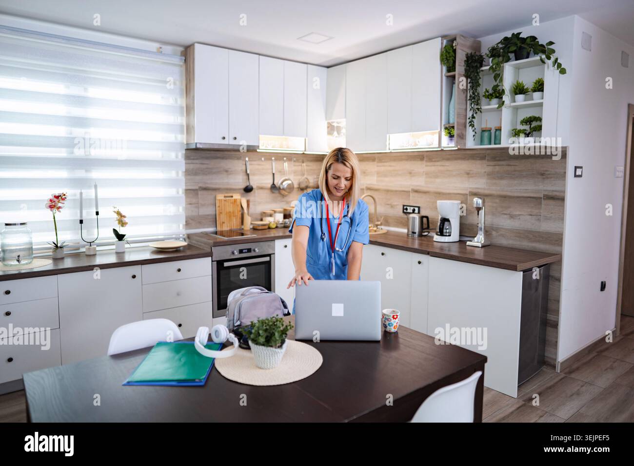 Eine Ärztin mit blauen Scrubs öffnet ihren Laptop in der Küche Stockfoto