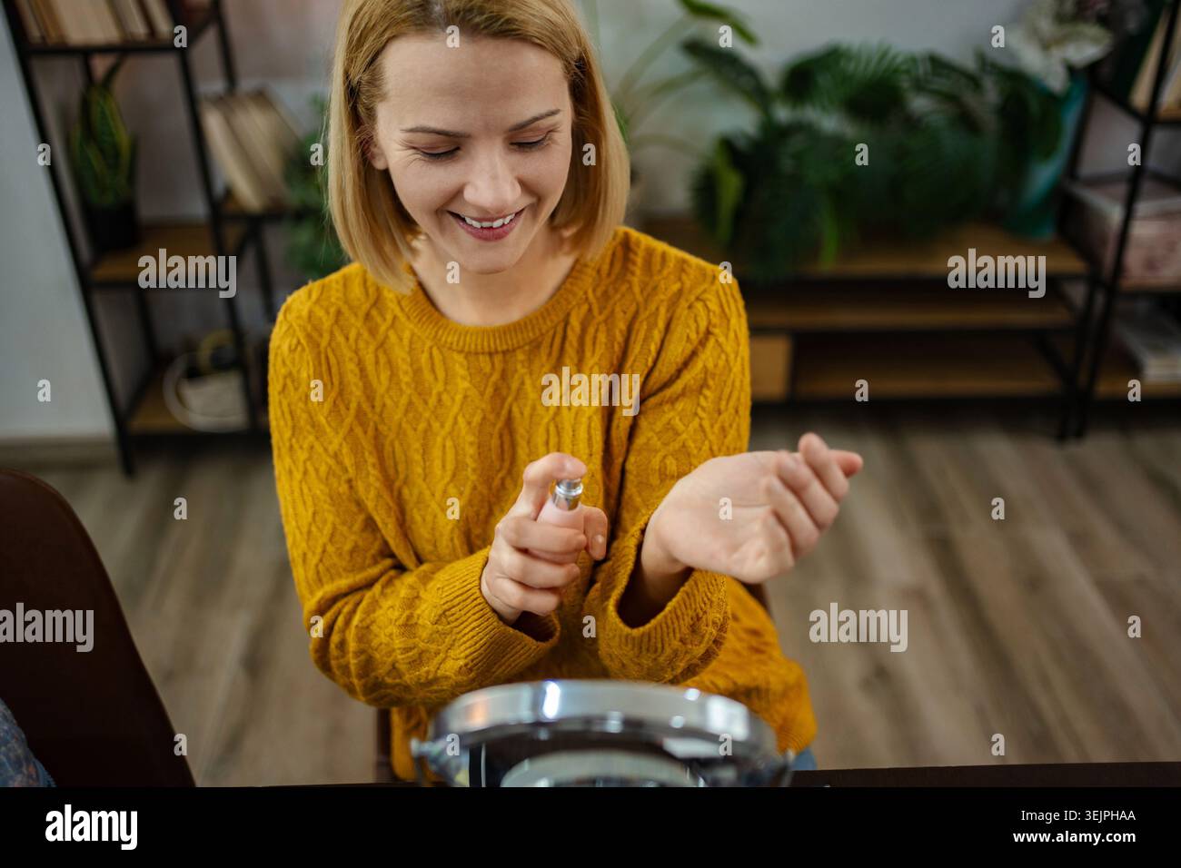 Bei einem Duftritual spritzt eine Frau Duft auf ihr Handgelenk, während ein fröhlicher Ausdruck ihr Gesicht in einem gemütlichen Ambiente erfüllt Stockfoto