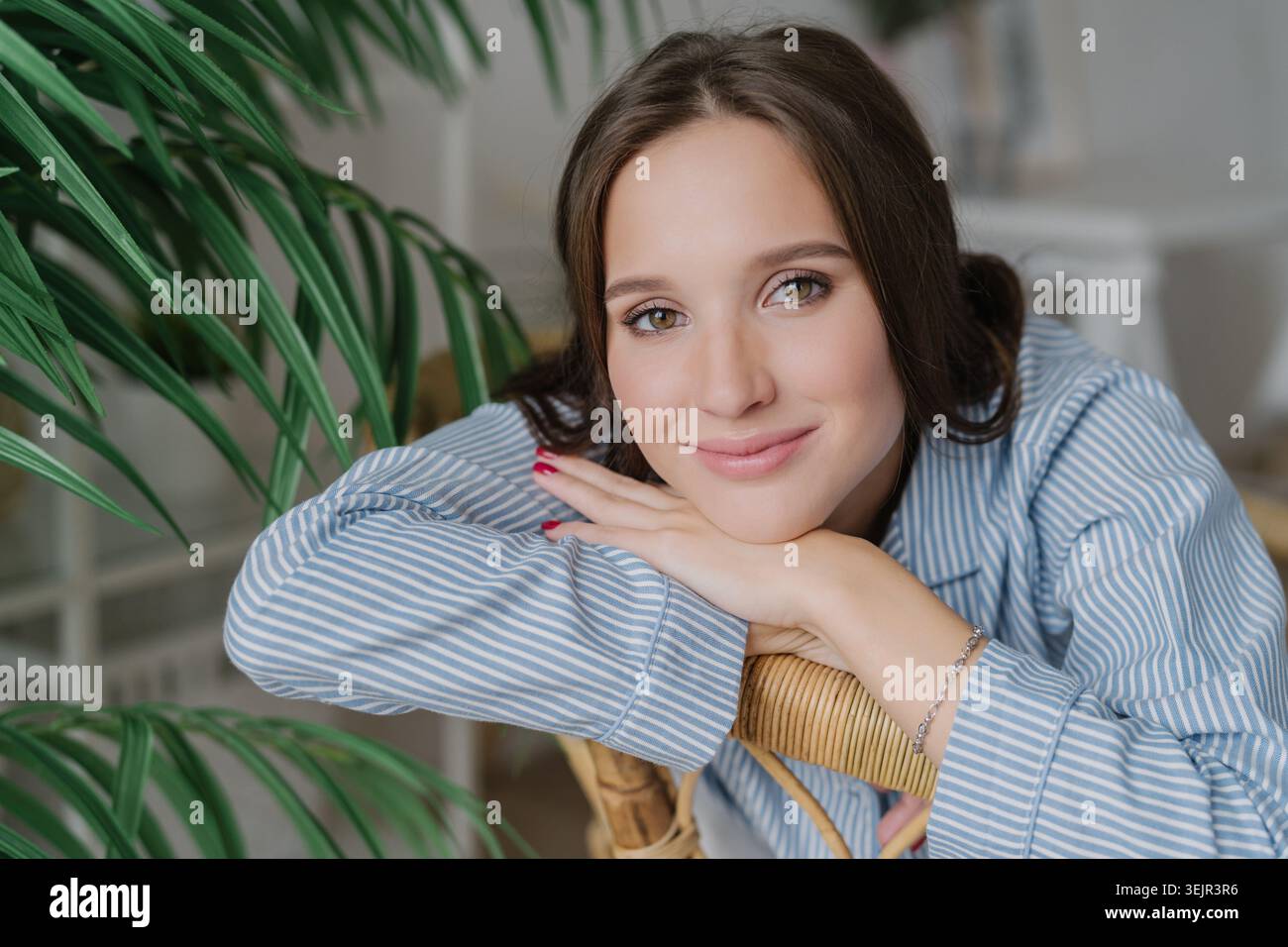 Nahaufnahme einer hübschen dunkelhaarigen jungen Frau hat ein charmantes, sanftes Lächeln, lehnt sich auf die Hände, posiert drinnen in der Nähe einer grünen Plantage, h Stockfoto