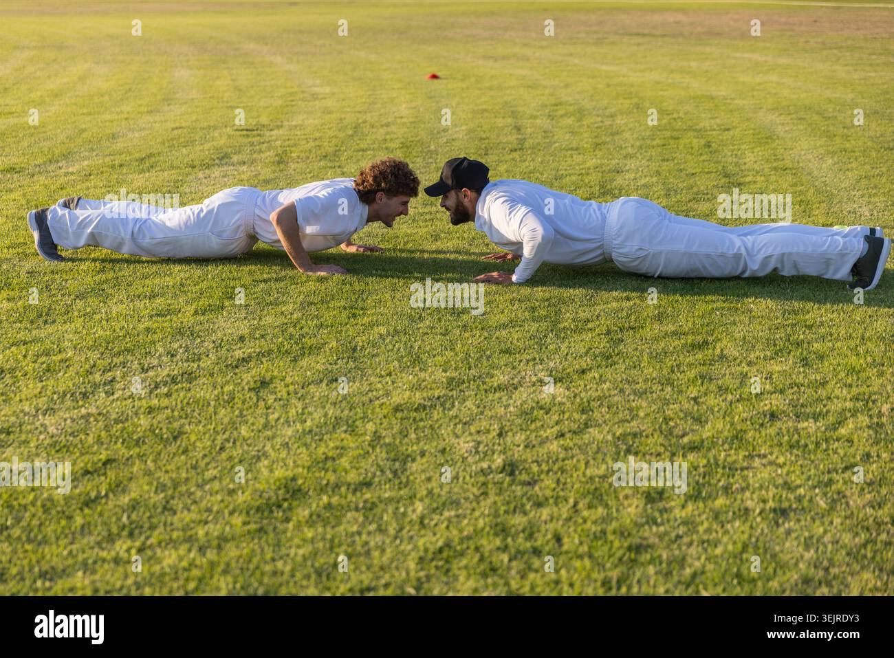 Zwei Männer, die Liegestütze auf dem Grasfeld in weißen Uniformen mit orangefarbenem Trainingskegel in der Nähe ausführen. Draußen, Rasen, Duo, sportlich, fi Stockfoto