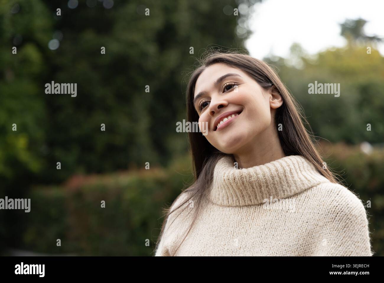 Eine Frau in ihren Zwanzigern lächelt, den Kopf neigt und im Park einen cremegestrickten Rollkragenpullover trägt. Porträt, draußen, Garten, Grün Stockfoto