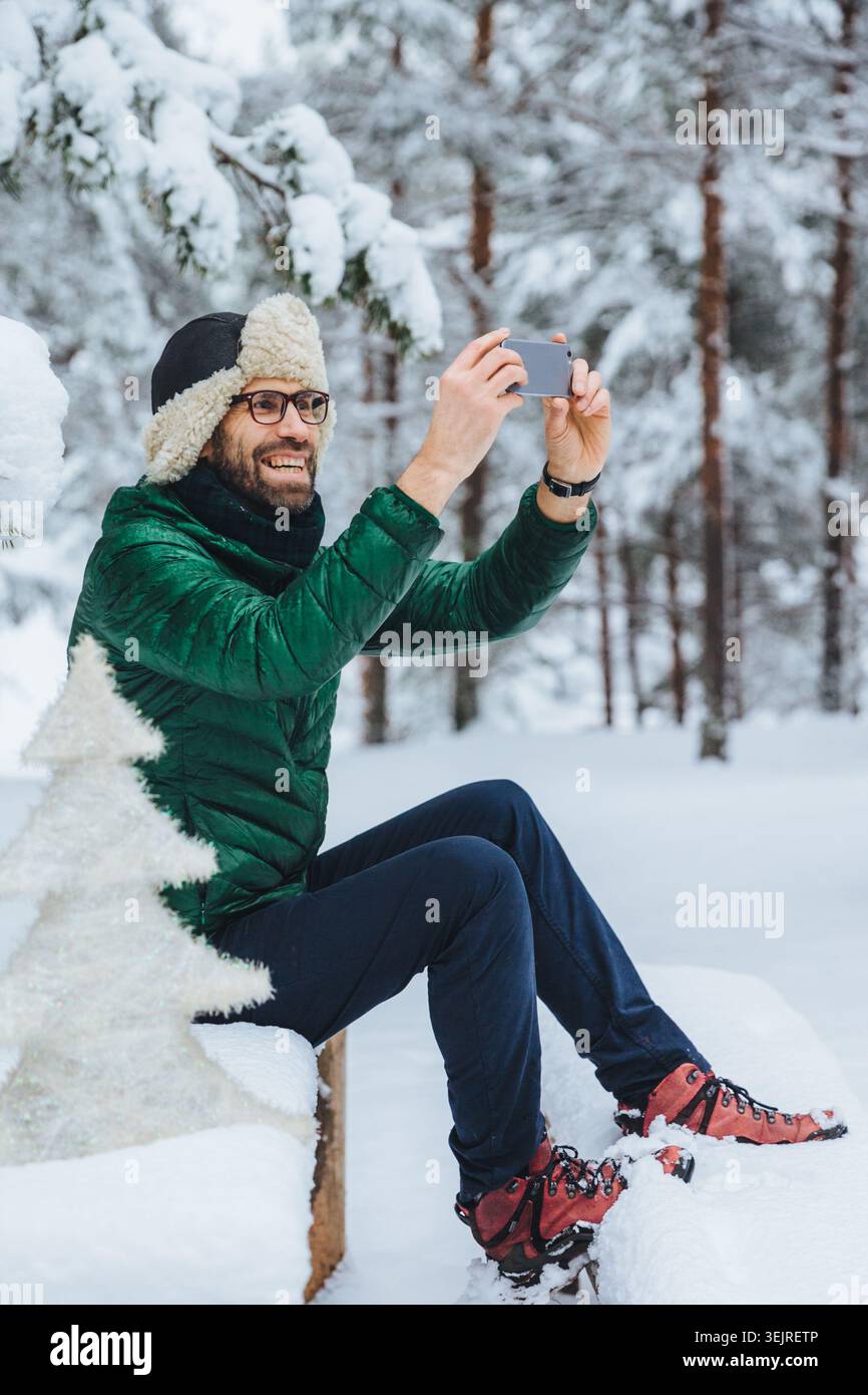 Ein bärtiger Mann benutzt das Smartphone, während er während der Winterwanderung im verschneiten Wald sitzt Stockfoto