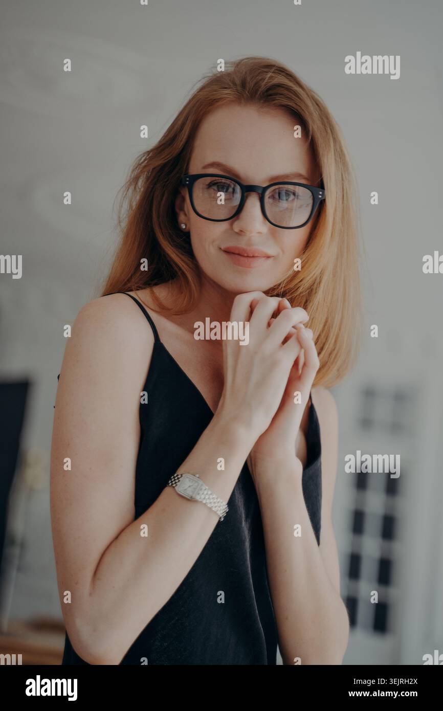 Rothaarige junge Frau, die eine schwarze Brille trägt, posiert drinnen mit Händen nahe dem Gesicht Stockfoto