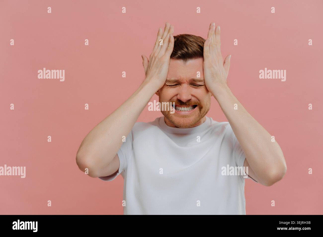 Gestresster Mann, der seinen Kopf mit beiden Händen gegen die rosa Studiowand hält Stockfoto