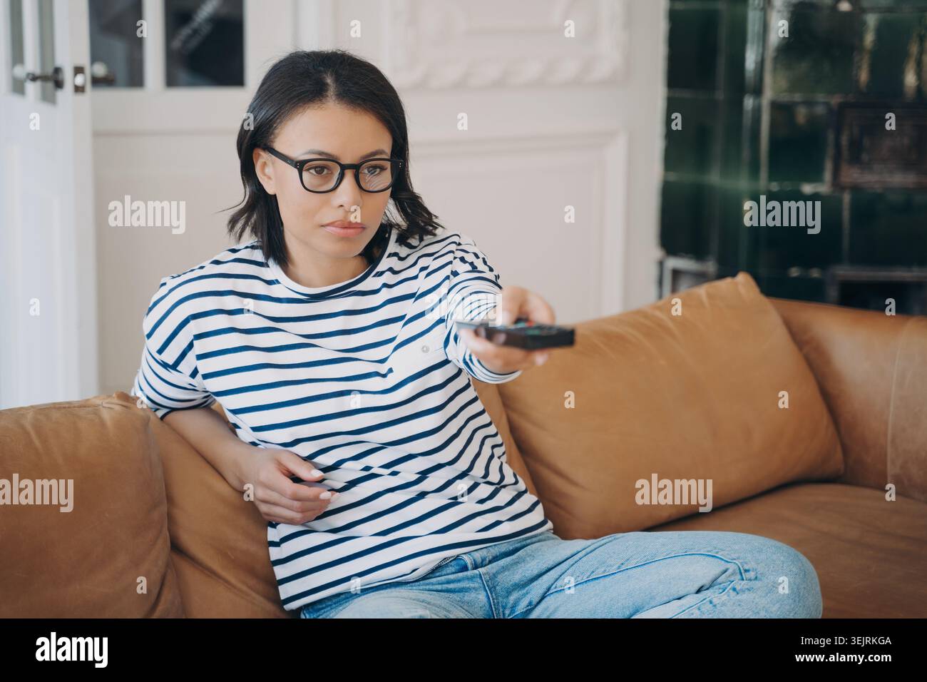 Eine junge Frau mit Brille benutzt die Fernbedienung, während sie sich auf dem Ledersofa im Wohnzimmer entspannt Stockfoto