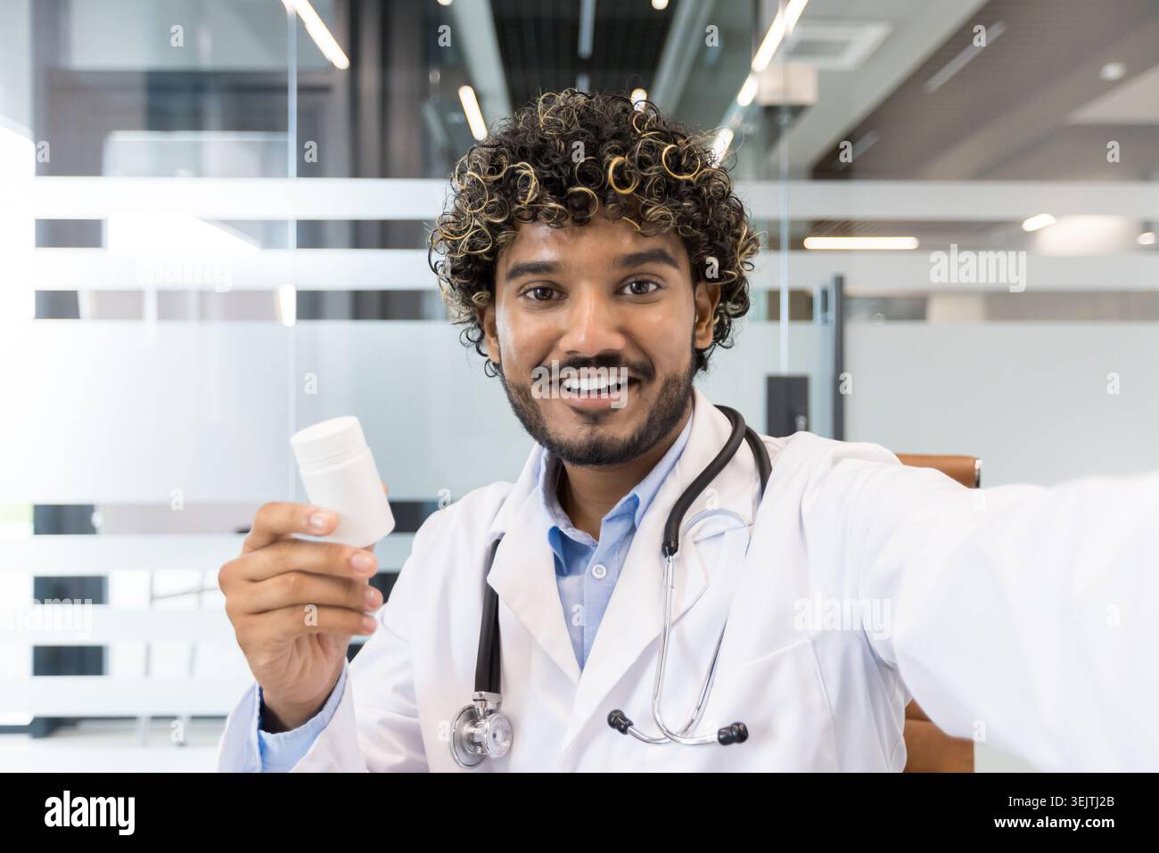 Junger indischer Arzt in einem weißen Labormantel und Stethoskop lächelt in die Kamera, hält eine Arzneiflasche, repräsentiert Telemedizin, Online-Gesundheitswesen und Remote-medizinische Beratung Stockfoto