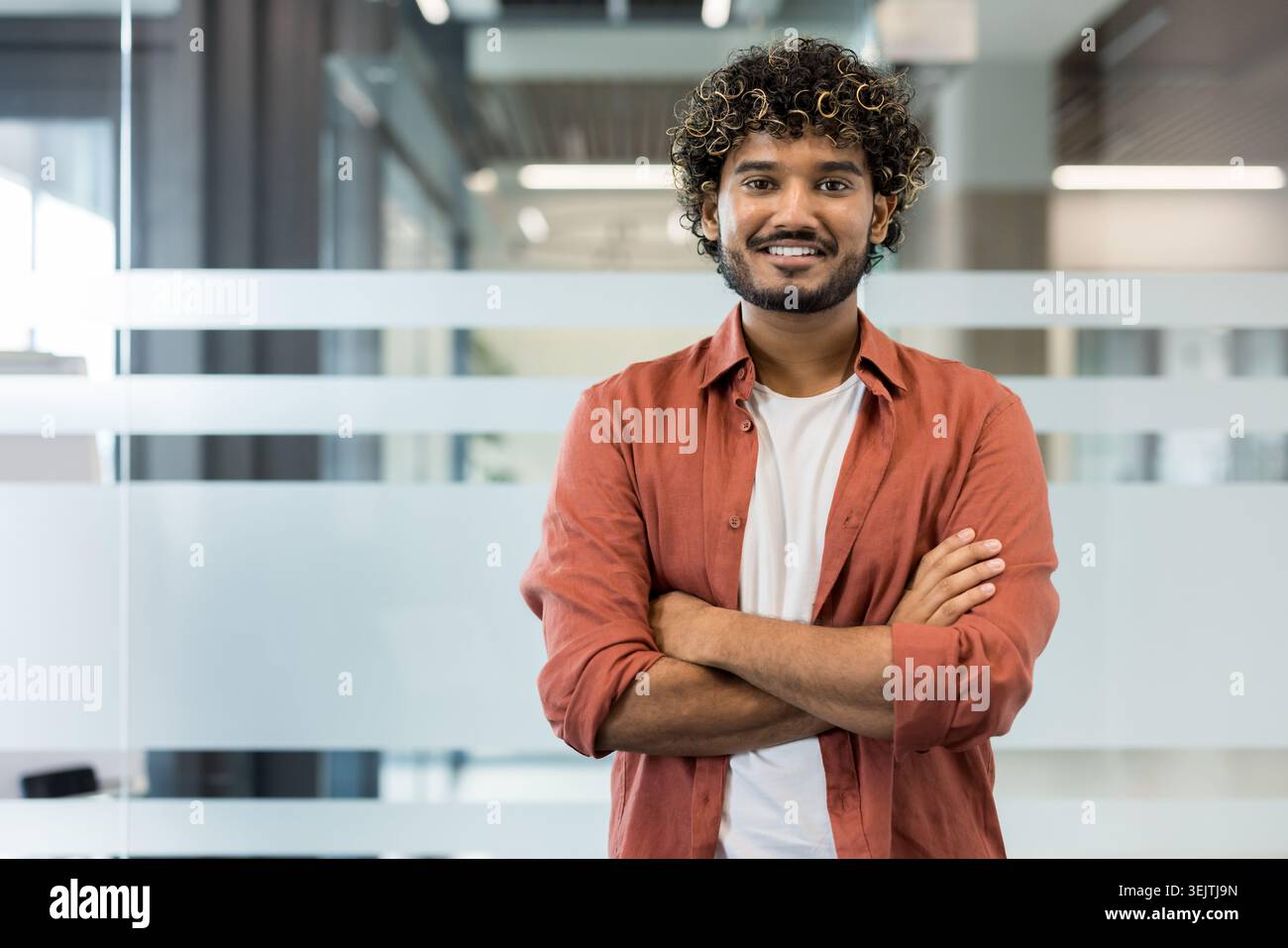 Junger, professioneller Mann mit lockigen Haaren und Bart, der in einem modernen Büro steht und selbstbewusst lächelt und die Arme überkreuzt. Er steht für eine freundliche und zugängliche Geschäftspräsenz Stockfoto