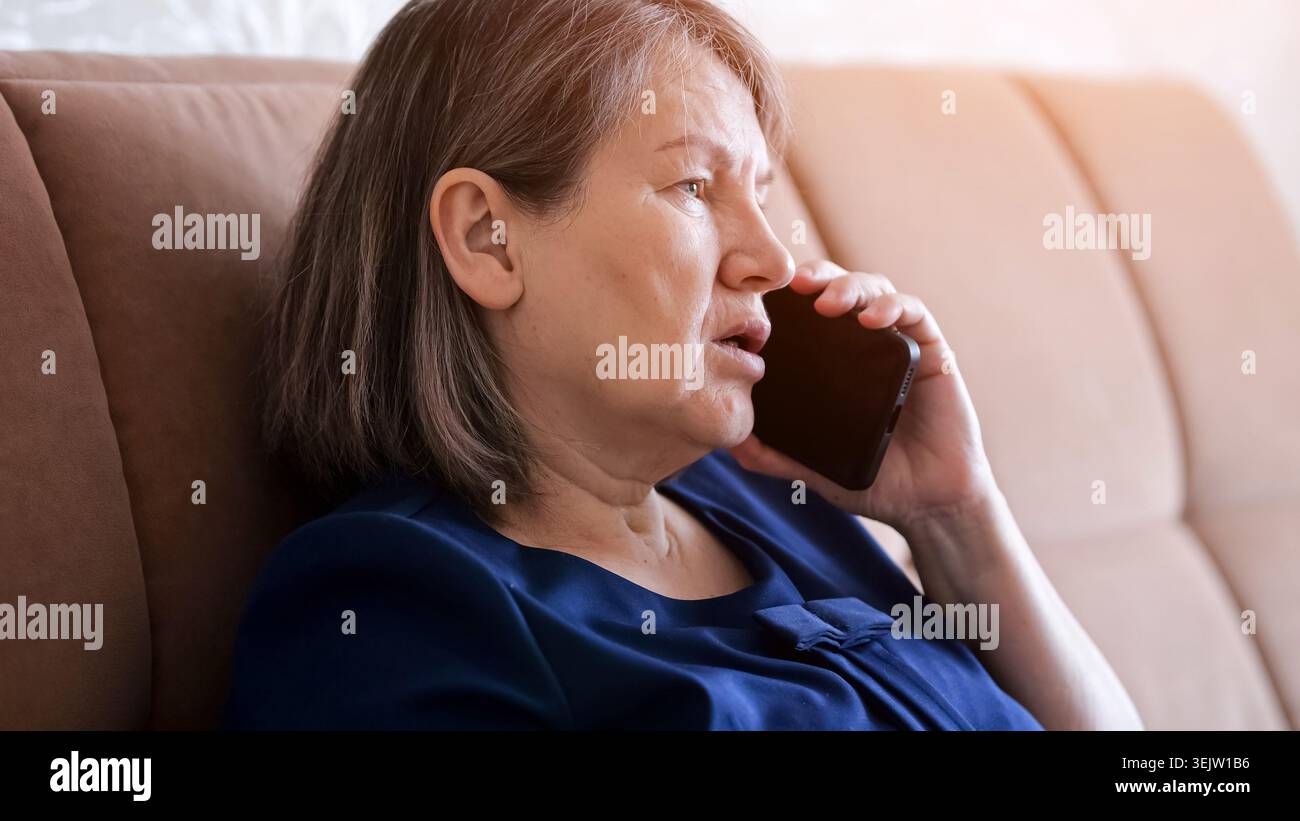 Ältere Frau zu Hause, die auf einem Sofa sitzt, ein Mobiltelefon am Ohr hält und mit besorgtem Ausdruck spricht, wichtige Nachrichten übermittelt oder Stockfoto