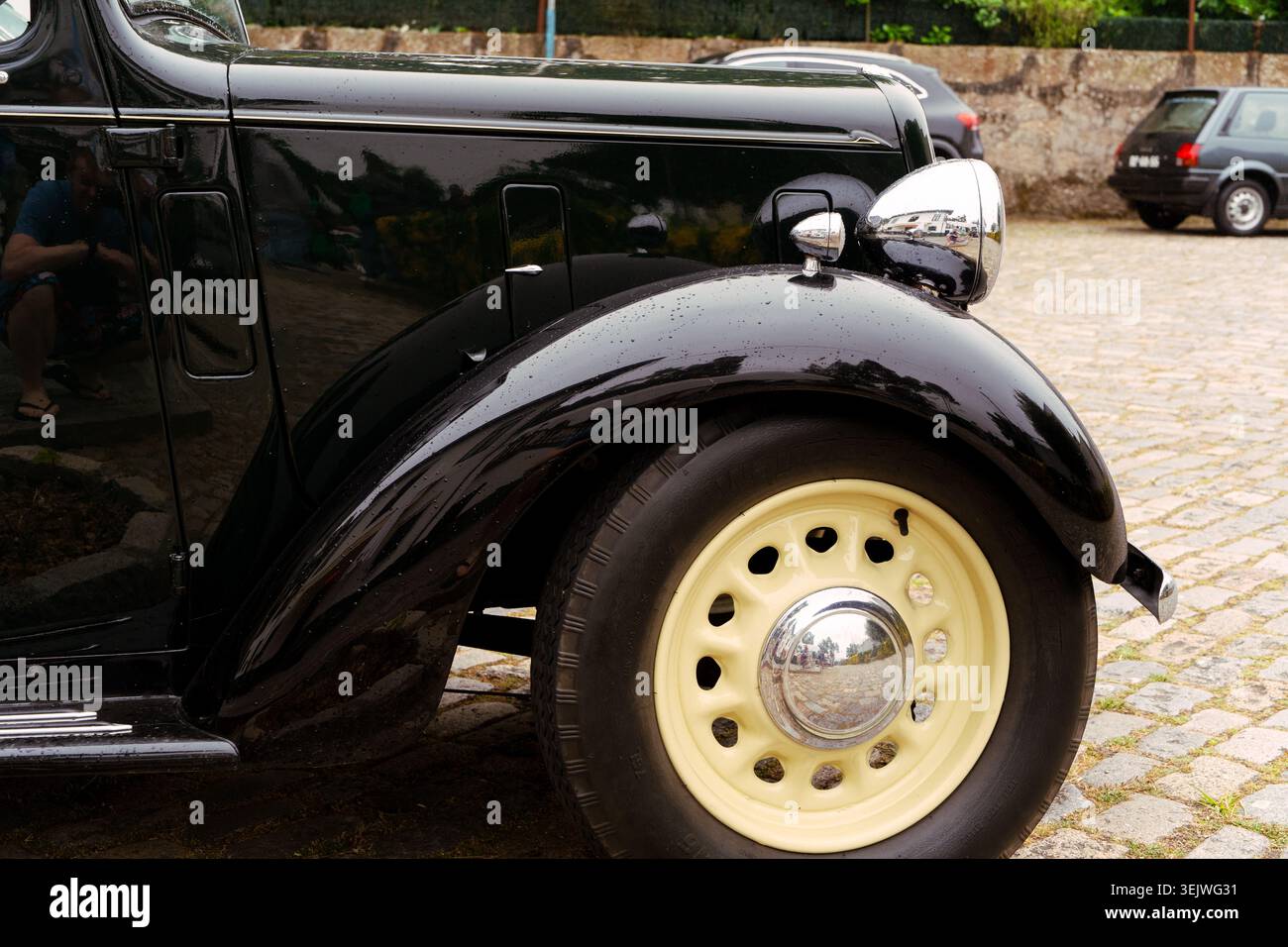Maia - Porto - Portugal , 30. Mai 2026: Schwarzer Oldtimer mit Chromscheinwerfer und cremefarbenem Rad auf Kopfsteinpflaster. Stockfoto