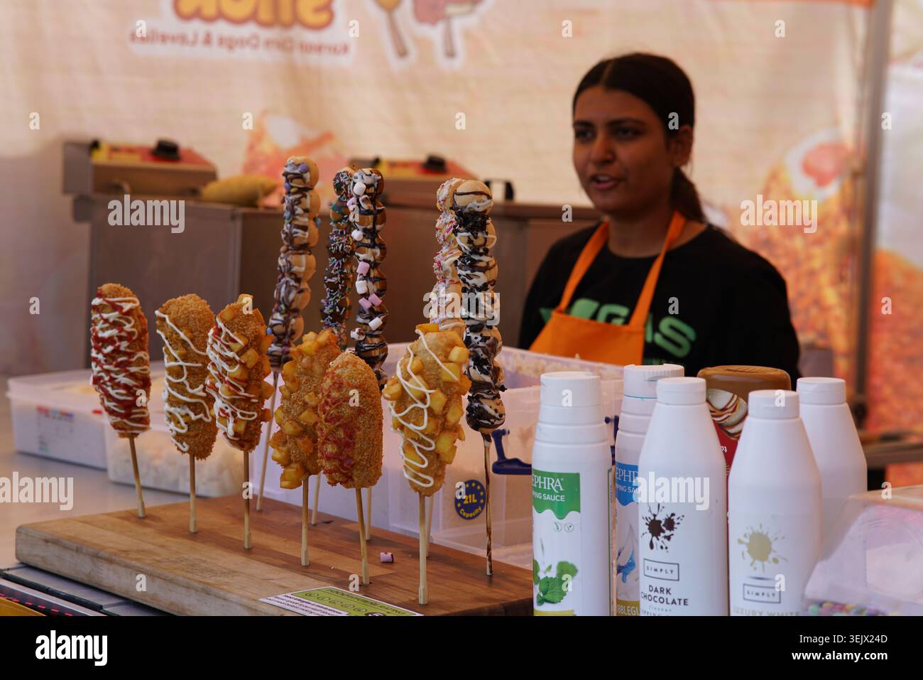 Die lächelnde Verkäuferin zeigt stolz verlockende, in Schokolade getauchte Obstspieße mit Erdbeeren und Bananen an einem lebhaften Marktstand im Freien. Stockfoto