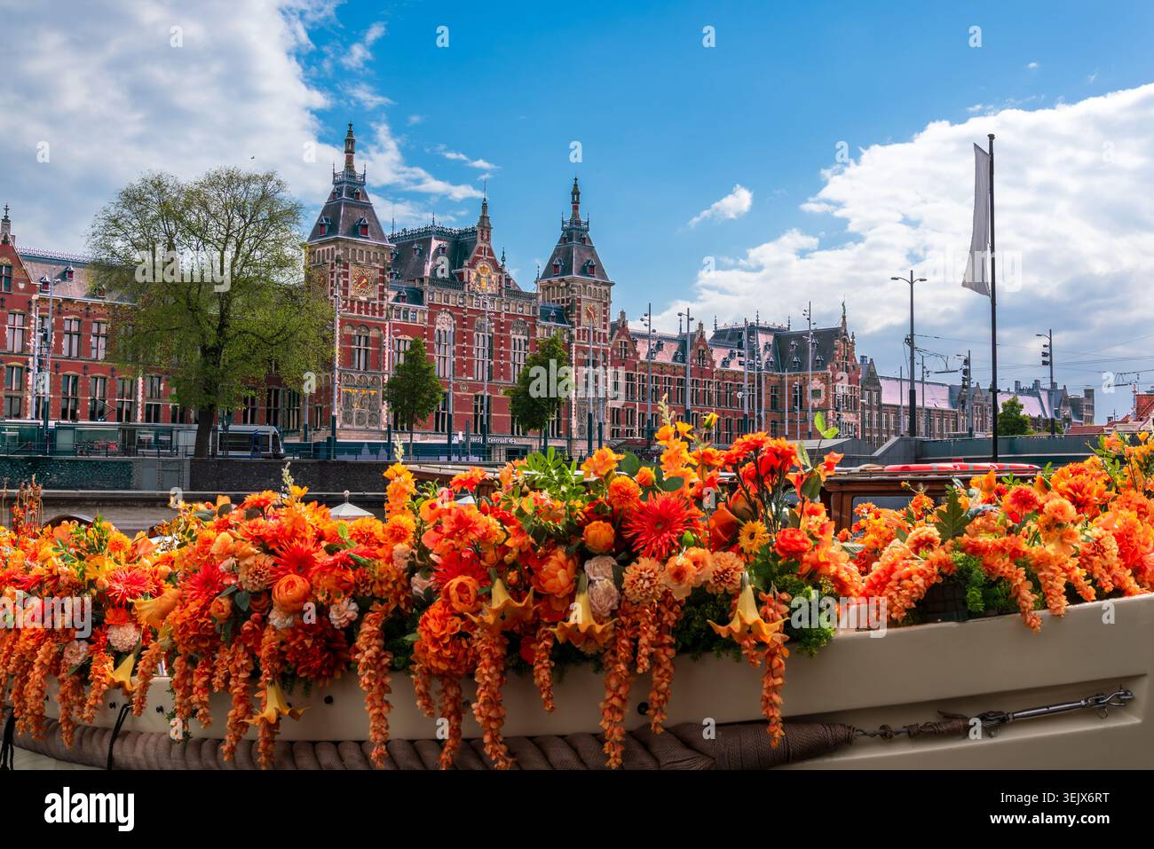 Amsterdamer Hauptbahnhof hinter leuchtenden orangefarbenen Blumen auf einem Kanalboot, historischer Bahnhof ohne Menschen, Niederlande Stockfoto