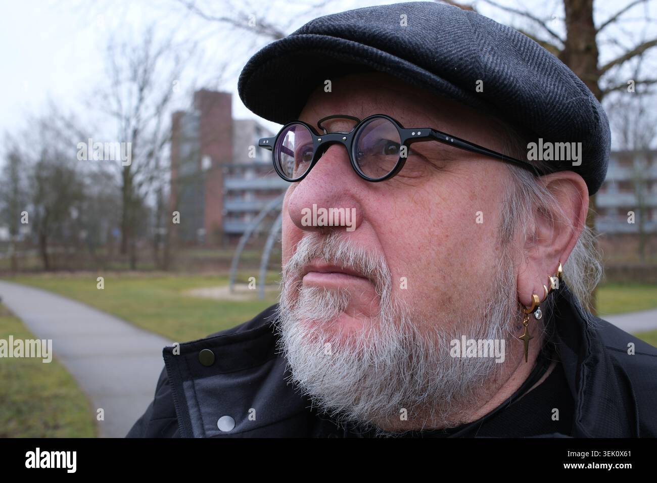 Ein älterer Mann mit grauem Bart und runder Brille, mit flacher Mütze und schwarzer Jacke. Mehrere Creolen und ein Kreuzohrring zieren sein linkes Ohr. Die Stockfoto