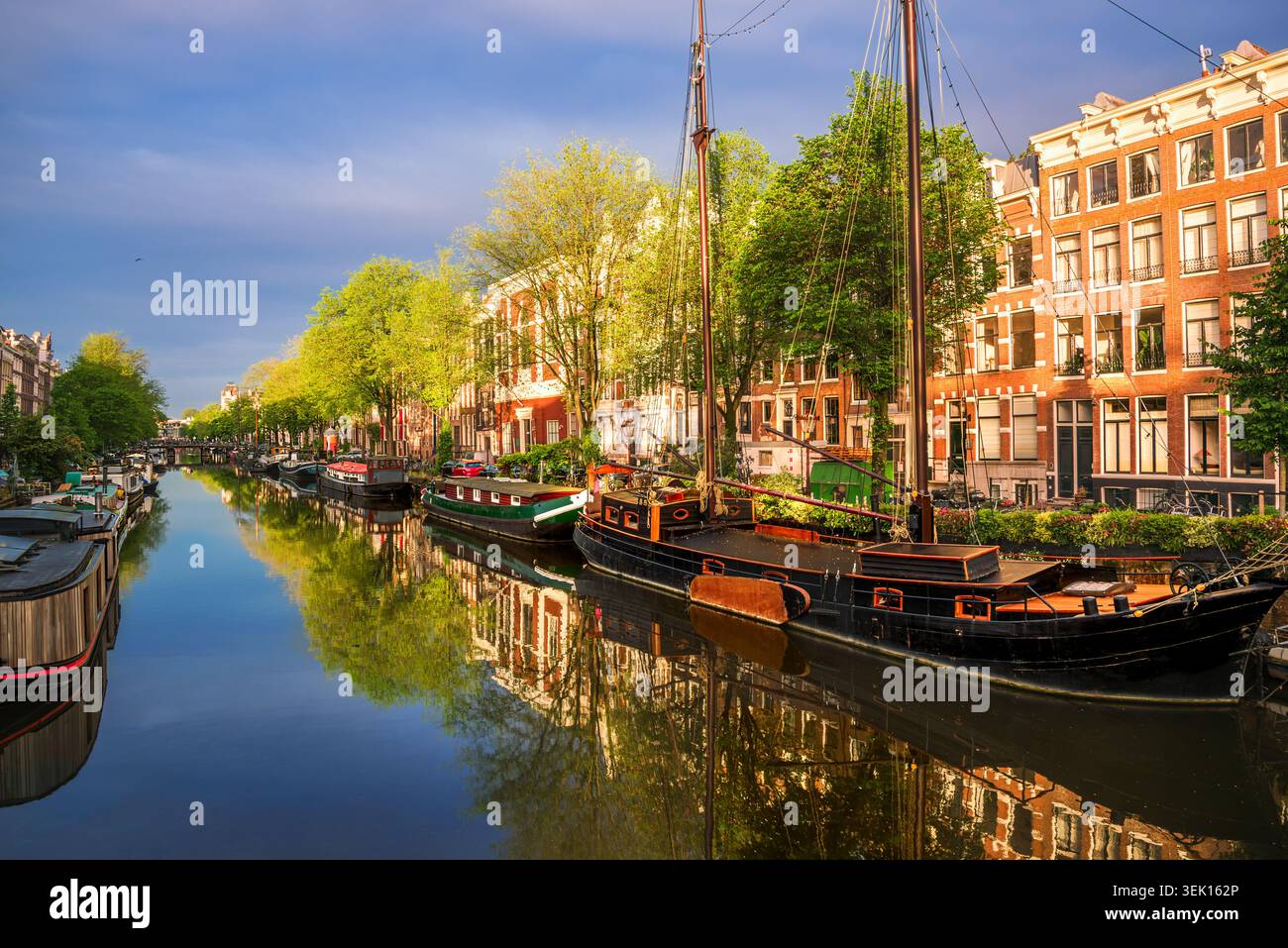 Traditionelle holländische Boote und historische Backsteinhäuser auf einem ruhigen Amsterdamer Kanal bei Sonnenaufgang, keine Menschen, Niederlande Stockfoto