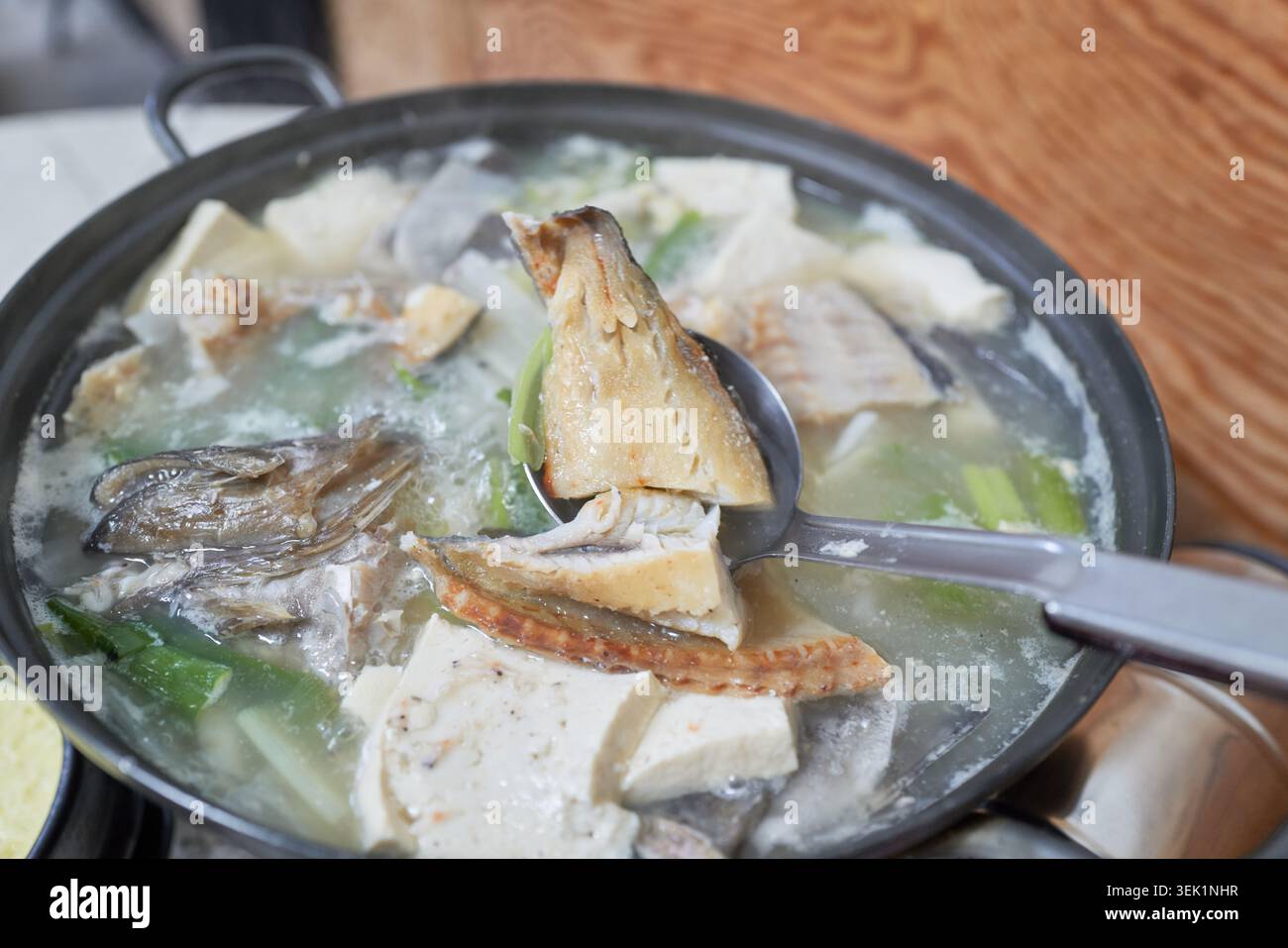 Koreanische halbgetrocknete Steinfischsuppe, Ureok-jeotguk. Eine Kelle hebt festen Fisch aus einer reichhaltigen Milchbrühe mit Tofu und grünen Zwiebeln in einem rustikalen dunklen Topf. Stockfoto