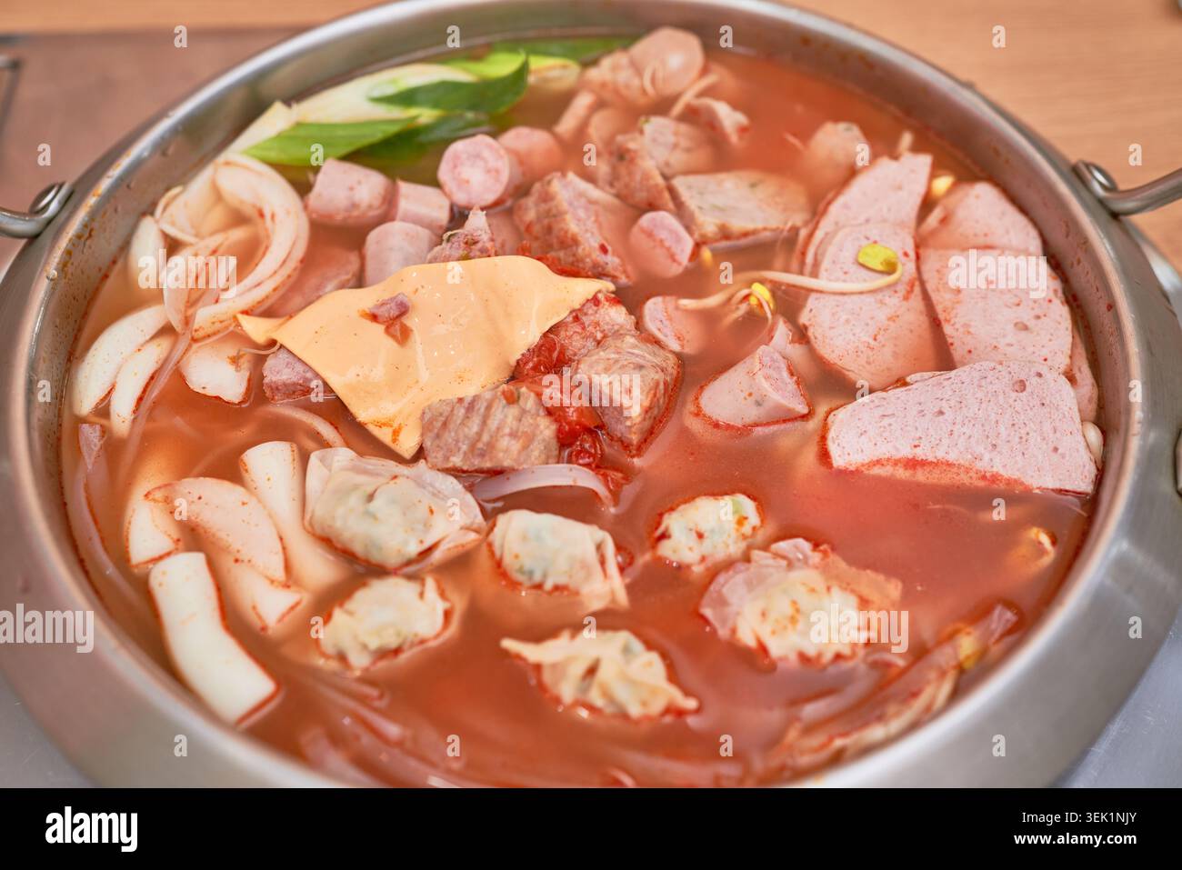 Koreanische Budae-jjigae in einem rostfreien Topf mit scharfer Brühe, gefüllt mit Würstchen, Spam, Käse, Knödeln und Ramen-Nudeln, die aktiv kochen. Stockfoto