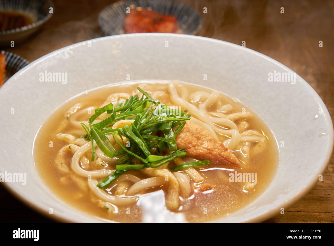 Eine Nahaufnahme von heißem Odeng Udon mit dicken Nudeln, in Scheiben geschnittenem Fischkuchen und grünen Zwiebeln in einer herzhaften Brühe, serviert mit einer Seite Kimchi. Stockfoto
