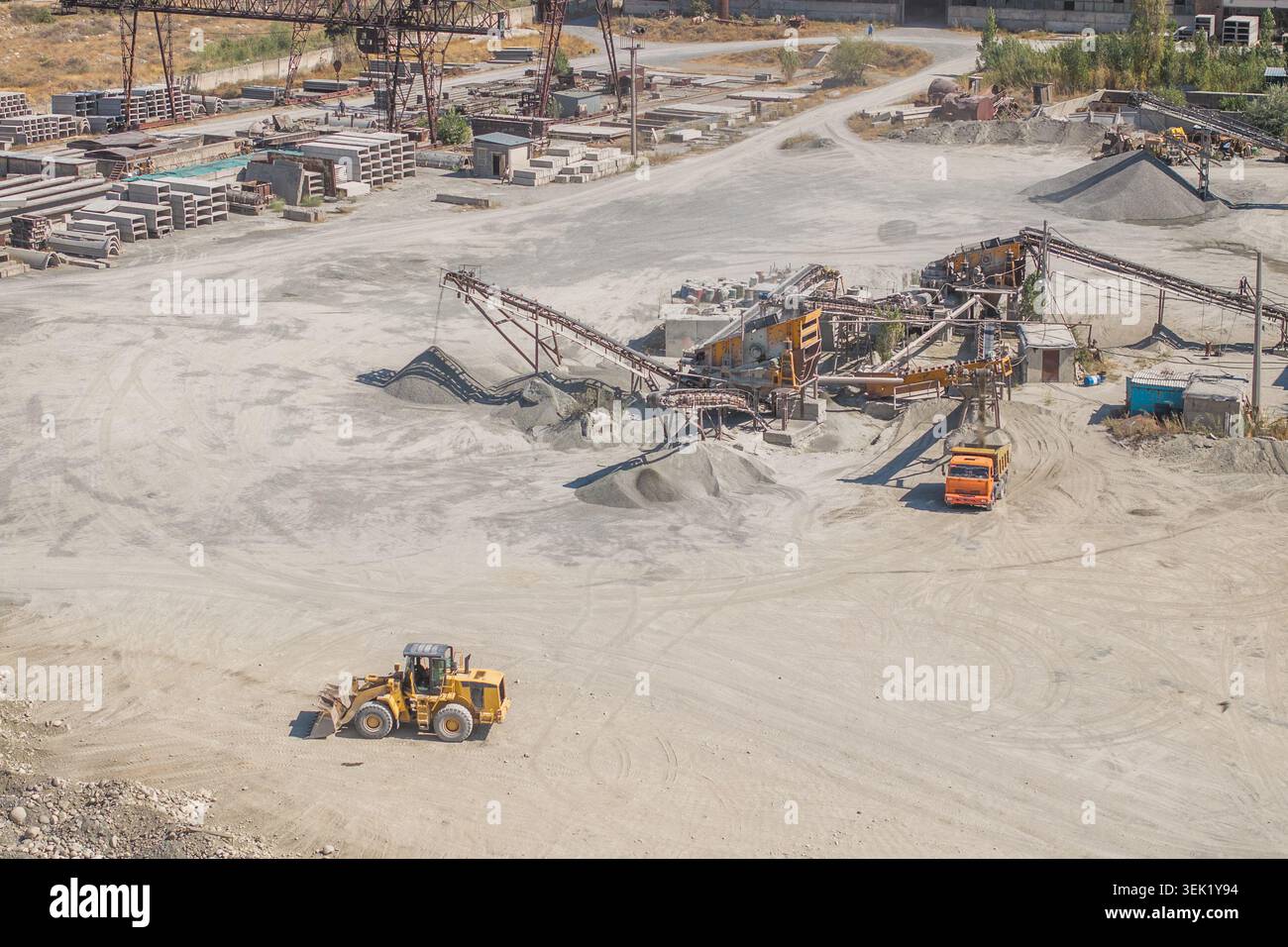 Die Luftaufnahme zeigt die geschäftige Industriebruche in Kirgisistan. Schwere Maschinen, darunter ein gelber Frontlader und ein orangefarbener Muldenkipper, werden in Kies- und Sandstapeln betrieben. Stockfoto