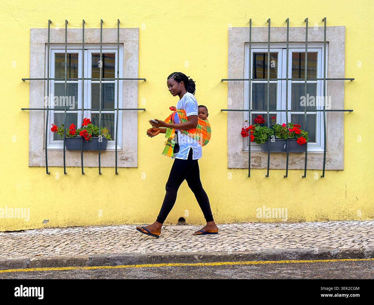 Mutter trägt Baby auf dem Rücken, gelbe Fassade, Alcântara, Lissabon, Portugal – lebendige Straßenfotografie Stockfoto