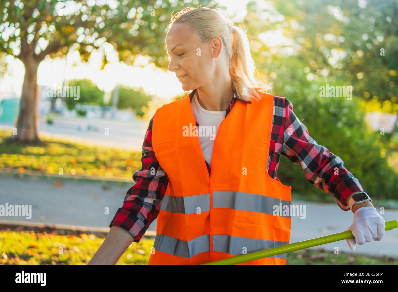 Frau in orangefarbener Sicherheitsweste und Handschuhen sammelt Müll in einem sonnigen öffentlichen Park und meldet sich freiwillig für die Gemeindereinigung, um die Gegend zu verschönern und zu schützen Stockfoto