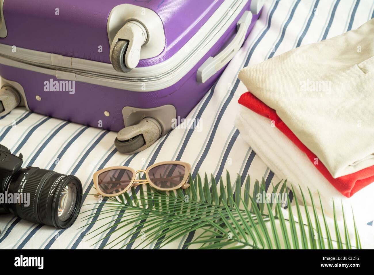Details aus violetten Kofferrädern, gefalteter Sommerkleidung und gestreiftem Bettzeug, die praktische Urlaubspackung zu Hause zeigen. Stockfoto