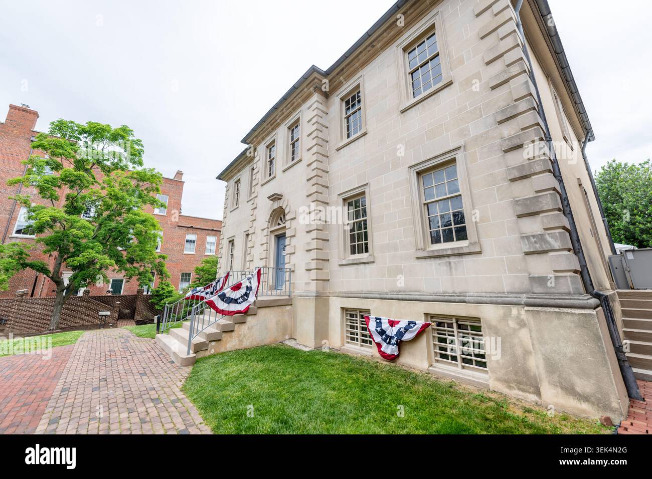 Carlyle House Georgische Steinfassade Altstadt Alexandria Vereinigte Staaten | ALEXANDRIA, Virginia — Patriotische Fahnen schmücken die Westfassade des Carlyle House in der Altstadt. Der 1753 vom schottischen Kaufmann John Carlyle fertiggestellte Herrenhaus wurde in rustiziertem Aquia-Creek-Sandstein erbaut — während der Restaurierung in den 1970er Jahren mit Indiana-Kalkstein neu verkleidet, um Verwitterung zu verhindern — und steht in scharfem Kontrast zur umgebenden Backsteinarchitektur. Die georgianische Struktur diente im April 1755 als Hauptquartier von Generalmajor Edward Braddock, wo er fünf kolonialen Gouverneure beim Kongress von Alexandria versammelte, um zu planen Stockfoto